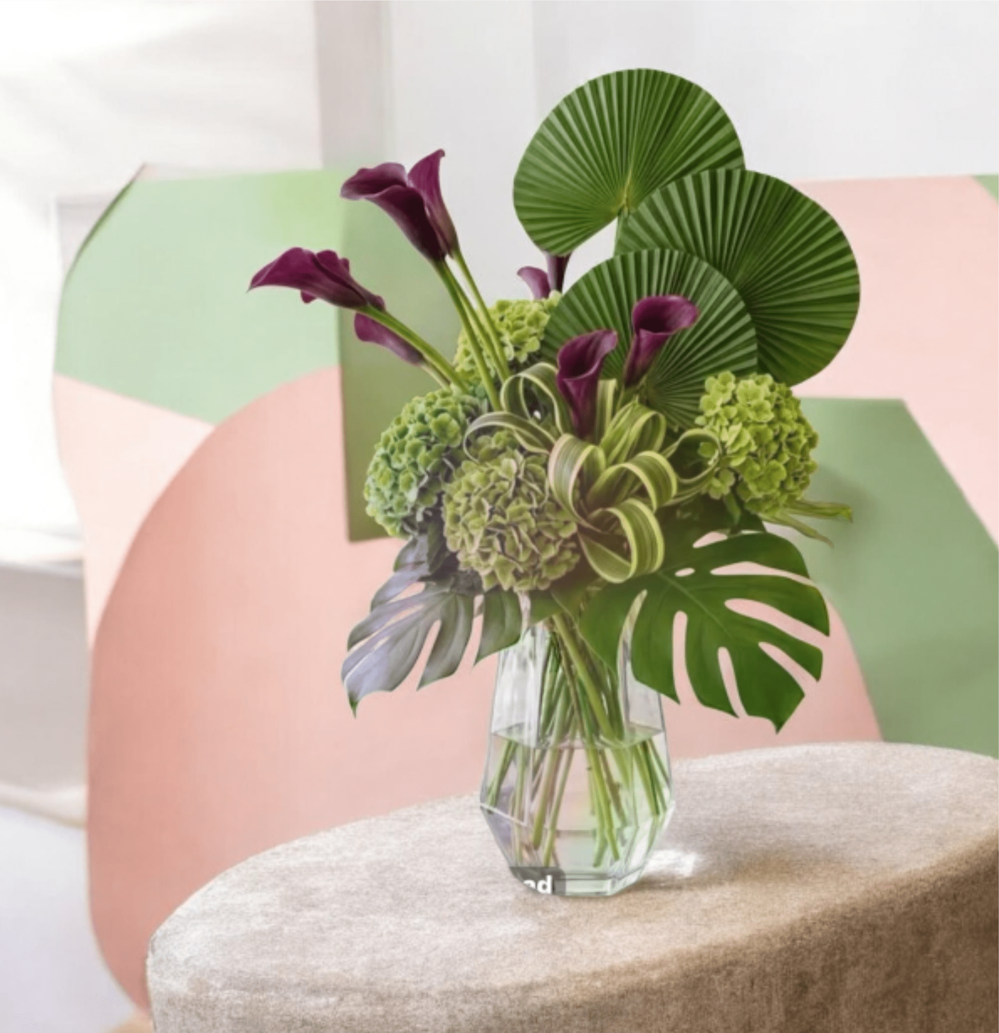 Armonia Purpura Tropical Flower Arrangement with Calla Lilies - Bright, tropical celebration bouquet designed to make the person you're thinking of feel truly seen on a birthday, housewarming, or congratulatory occasion. Towering pink hibiscus-like blossoms and fiery orange bird-of-paradise spikes pair with sunny yellow lilies for an instant burst of warmth and cheer; glossy palm fronds and layered greenery create an upright, sculptural silhouette that reads medium-to-tall and makes a bold entrance.  This arrangement mixes textured shapes-rounded red roses and clustered green hydrangeas against tall purple calla lilies and smaller orange and white accents-for striking contrast in both color and form. Presented either in a natural woven basket for a relaxed, casual vibe or a black rectangular vase for a modern, structured statement (one version uses a clear glass vase to showcase the stems), the palette of pink, yellow, orange, red, purple and fresh green stays bright and celebratory. Materials include lilies, roses, carnations, calla lilies, hydrangeas and tropical blooms with decorative palm and monstera-style leaves for added drama.  Choose this when you want the recipient to feel celebrated-perfect for tabletops, entryways, or anywhere you want an immediate lift of color and texture. The upright profile ensures strong presence; order with local delivery for best freshness and immediate impact.