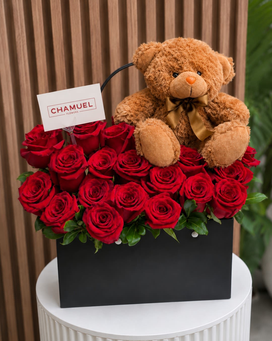 Candelaria Red Rose Box Arrangement with Teddy Bear - A compact cluster of vivid red roses set against a matte black gift box creates a bold, romantic statement-perfect as a birthday bouquet, anniversary roses, or a Valentine's Day surprise. The Candelaria boxed arrangement reads as a premium, love-themed gift: the deep red blooms pop with passion next to black accents, and the included plush teddy bear with a satin bow softens the look so the recipient feels both adored and cherished.  This arrangement features densely packed red roses tucked into a sleek black gift box with a ribbon, branded card, and a convenient handle for easy presentation and transport. The elevated, compact design makes it ideal for delivering a heartfelt message without needing a vase; the plush bear adds a playful, affectionate touch that complements the roses' classic romance. Choose this for someone you want to make feel truly seen-whether you're marking an anniversary, saying "I love you," or celebrating a special day.
