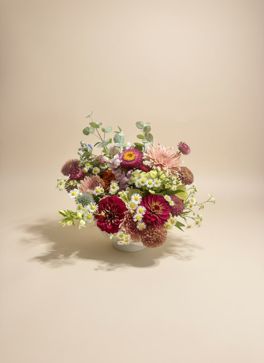 Vintage Centerpiece  - A compact, rounded flower arrangement that feels like a pocket of summer-perfect as a birthday,, thank-you gift, or just-because surprise to make someone feel seen. The pink, white, purple, and green palette reads fresh and cheerful, so the person you're sending it to will notice the soft, lively mix of color right away.  Arranged in a low white ceramic bowl vase, this composition pairs small daisy-like blooms with larger chrysanthemum-style heads and airy eucalyptus stems that add gentle height and texture. The garden-style layering of blush, magenta, and white petals against fresh greenery creates a full, natural look that sits neatly on tabletops or desks. Sized for easy display and effortless charm, it's an inviting choice for spring occasions or everyday celebrations.  Please not all of our designs are custom and we use what is available day off. So flowers and colors might vary. 