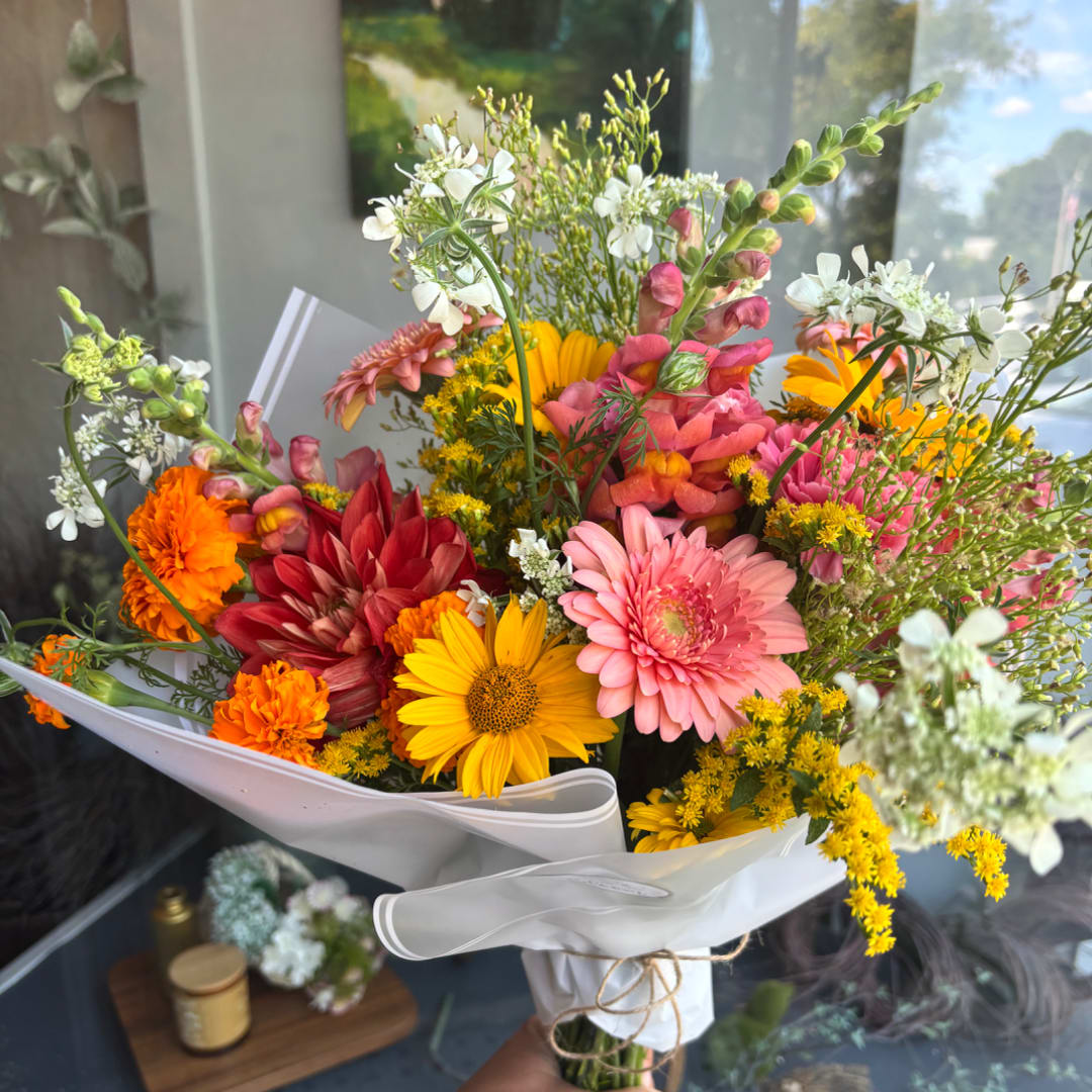 Handheld bouquet of mixed pink, yellow, orange, and white flowers wrapped in white paper