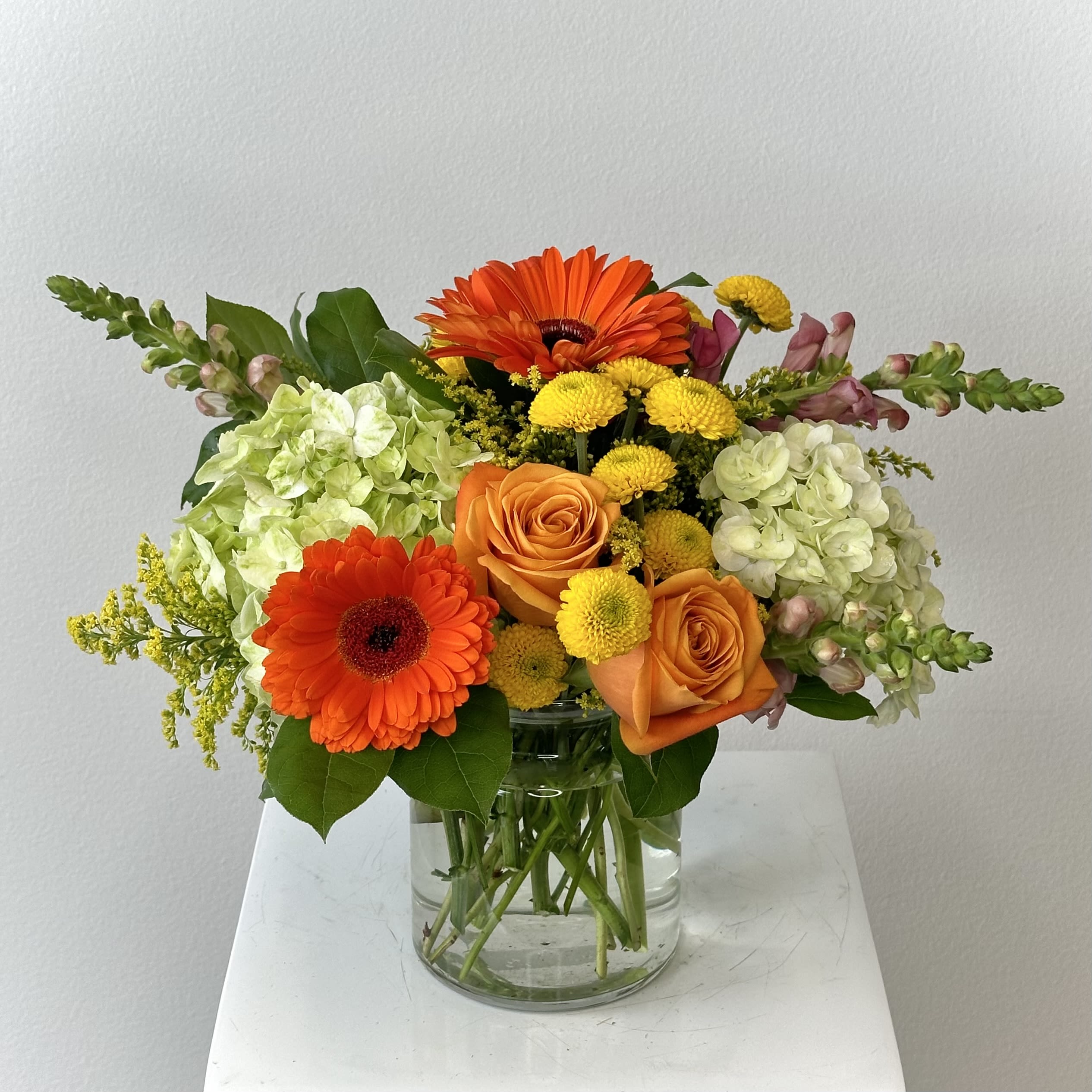 Orange gerbera daisies and roses in a clear glass vase with pale hydrangeas