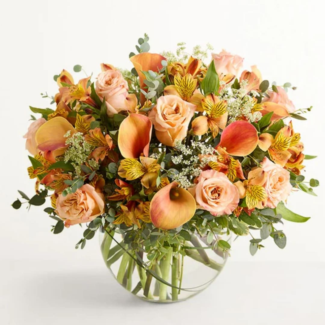 Graceful Fall  - Soft peach roses and vibrant orange calla lilies mingle with sunny yellow-striped alstroemeria in a graceful fall flower arrangement perfect for a birthday bouquet, thank-you gift, or a springtime celebration. The rounded clear glass vase keeps the look modern and clean while showing the green stems below, so the recipient will feel instantly uplifted by the bright, cheerful palette that still reads gentle and romantic.  This arrangement includes peach roses, mini calla lilies and full-size calla lilies, alstroemeria with yellow striping, small white filler blooms, and airy eucalyptus-like foliage for texture and movement. The mix of peach, orange, yellow, green, and soft pink tones creates a fresh, versatile display that suits birthdays, anniversaries, or just-because moments.  Ideal for the person you want to make feel seen and celebrated, Graceful Fall sends a warm, sunny message without being overly formal. Delivered in a clear round glass vase, it arrives ready to display; change the water every few days, trim stems slightly, and keep away from direct heat to extend the life of the blooms.