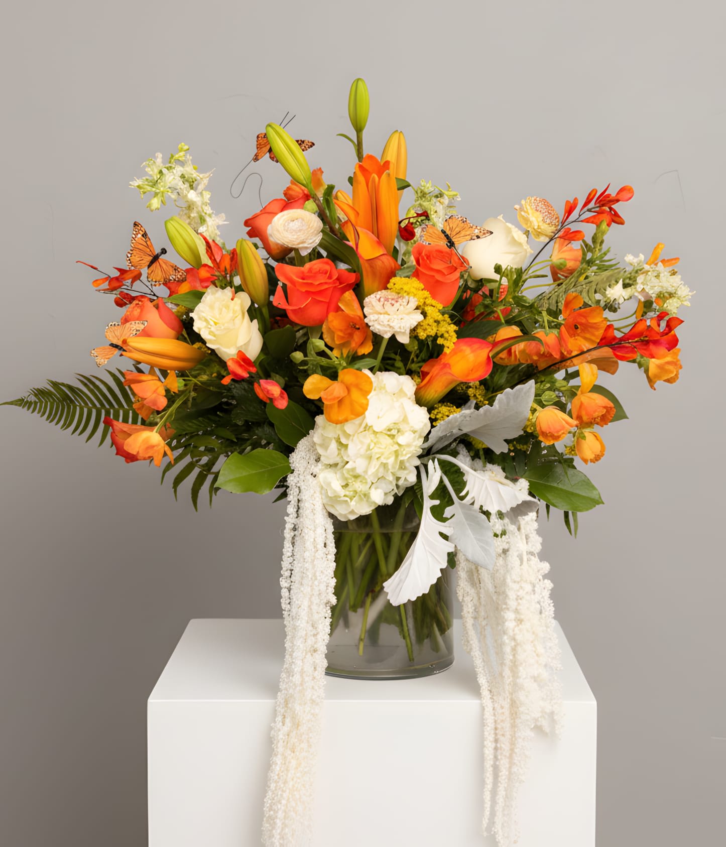 Autumn Butterfly Symphony - Bold, artistic, and full of movement, Autumn Butterfly Symphony bursts with vibrant orange lilies, roses, calla lilies, freesia, hydrangea, and seasonal textural blooms accented with delicate butterfly details and cascading white amaranthus. Designed in a modern clear cylinder vase, this luxurious statement arrangement captures the warmth and beauty of a glowing autumn garden in full bloom. Perfect for grand celebrations, upscale events, birthdays, or unforgettable centerpieces.