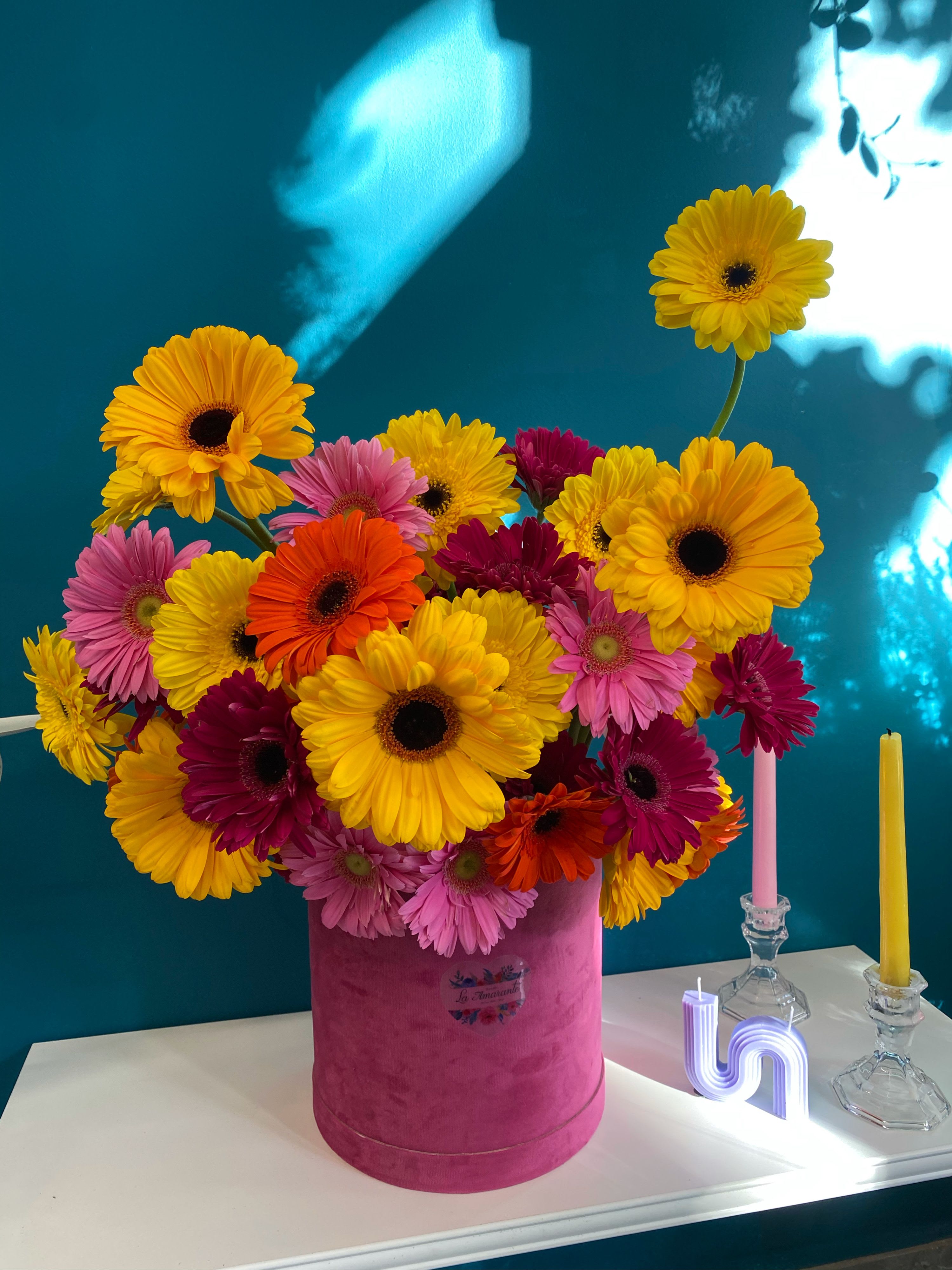 Bright gerbera daisy arrangement in a pink hatbox with candles nearby