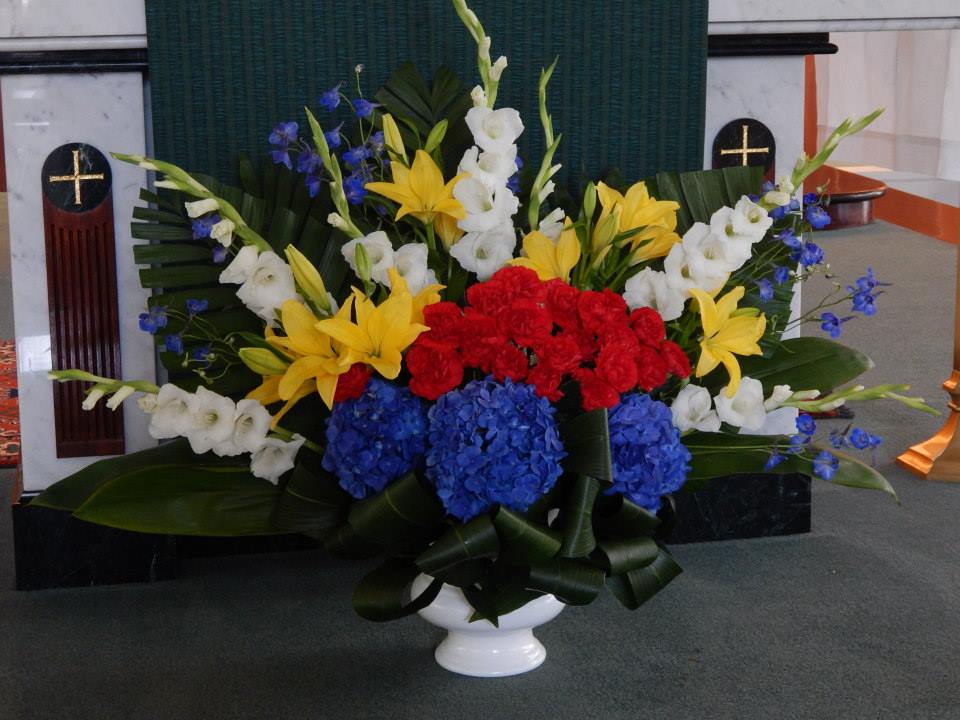 Large funeral arrangement with red, yellow, white, and blue flowers in a white vase