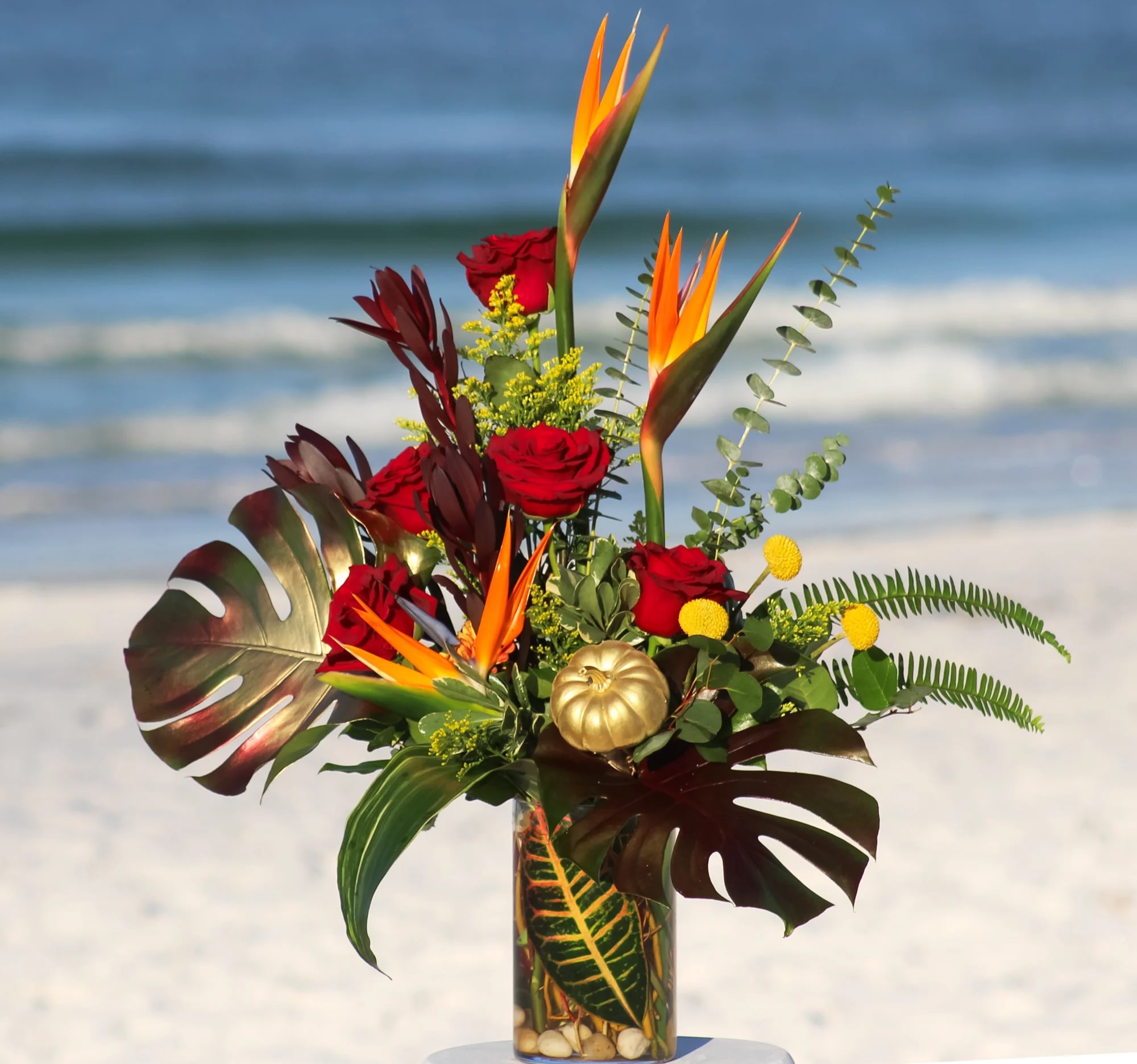 Roses &amp; Paradise - This beautiful unique design includes red roses, birds of paradise, Safari Sunset, crespedia, yellow solidago, with a gold mini pumpkin. Lush tropical foliage such as large monstera leaves tinted in gold and burgundy, baby blue eucalyptus, sword fern, aspidistra leaves, and a croton leaf inside the vase. 