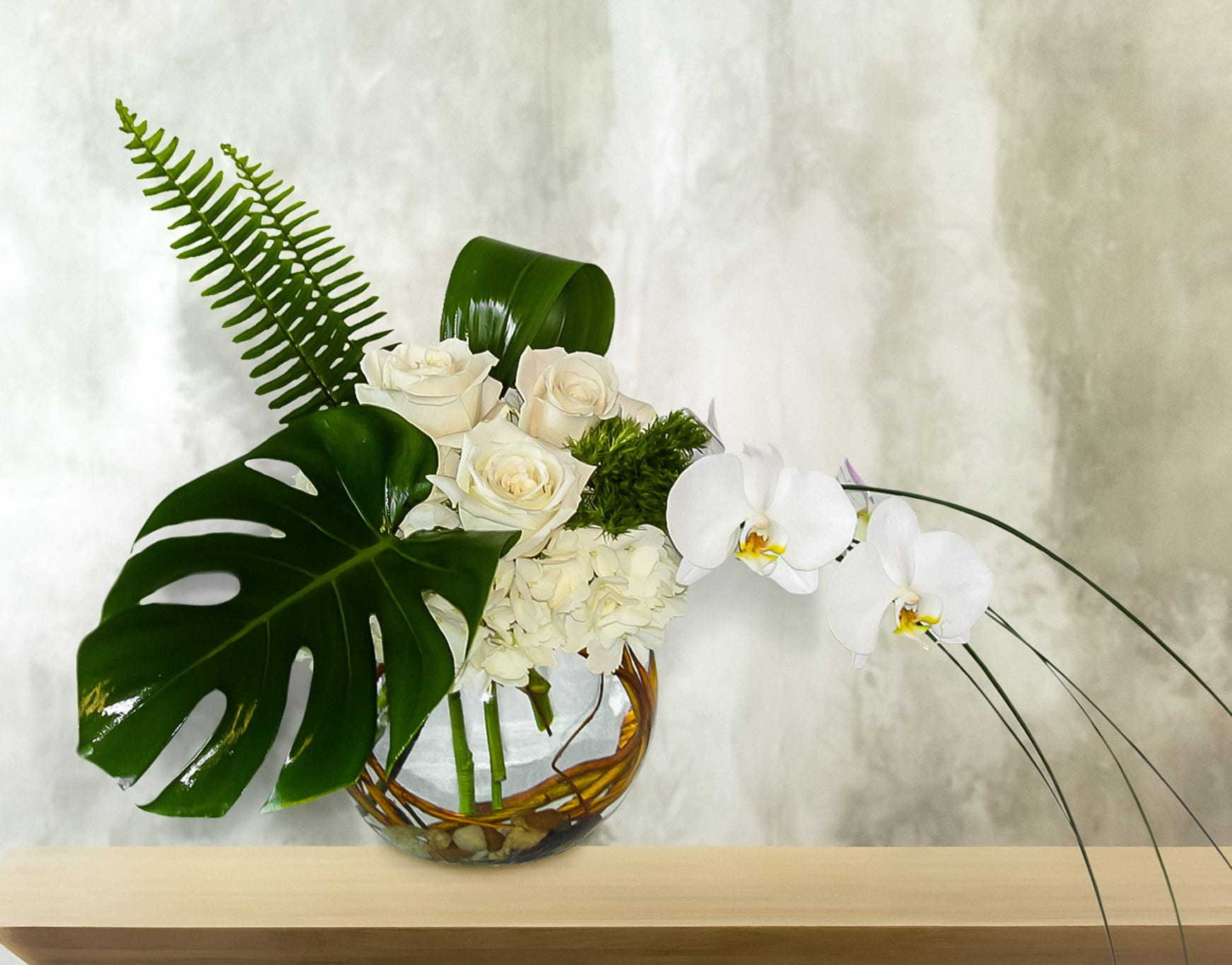 White roses and orchids arranged in a glass bowl with large tropical leaves