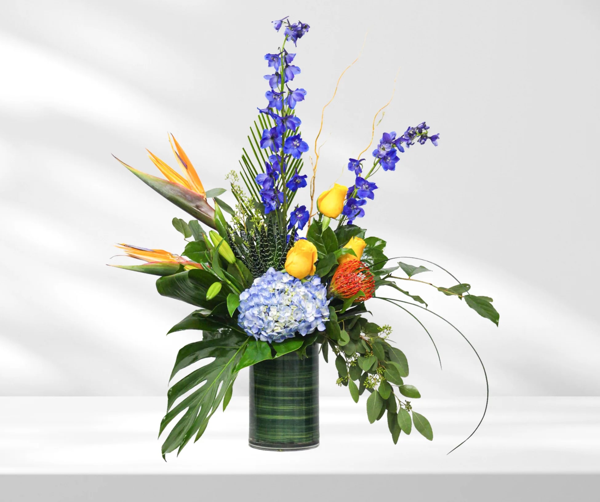Tropical arrangement with blue flowers, yellow roses, and bird of paradise in a glass vase