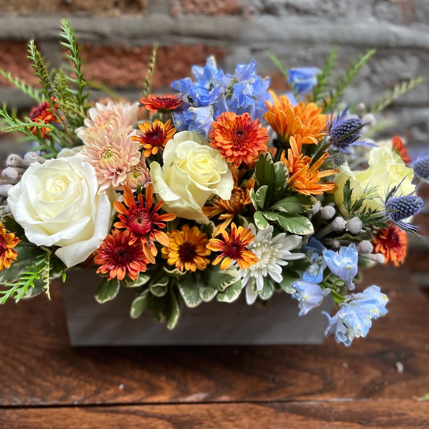 Mixed bouquet with white roses, orange daisies, and blue flowers in a rectangular container
