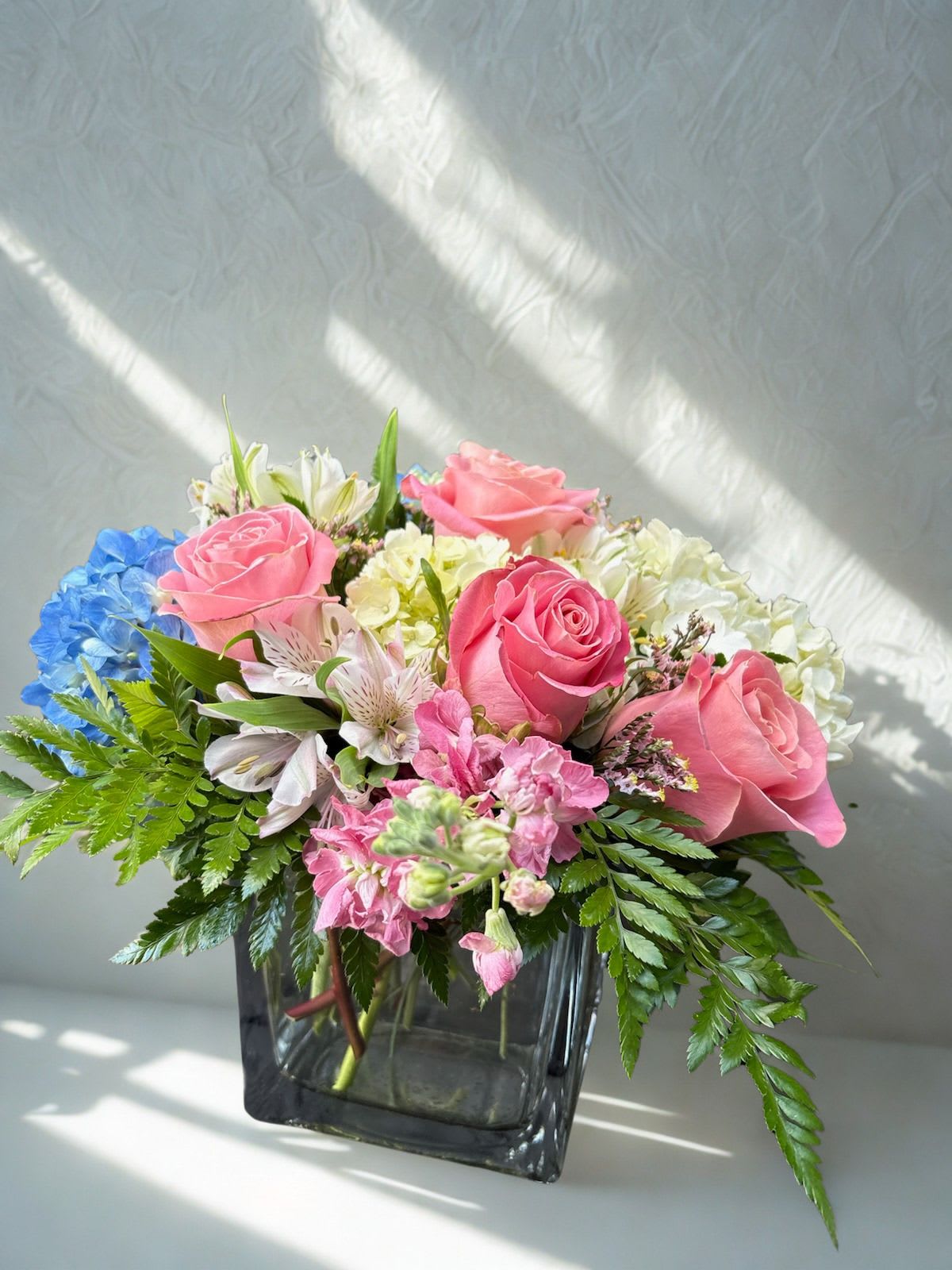Its A ....??? - Soft, cheerful, and romantic, this mixed bouquet of pink, white, and blue blooms feels like a warm embrace, making it the perfect gift for birthdays, gender reveal,  anniversaries, or simply to say "I'm thinking of you." The arrangement features a low, spilling composition of clustered pink roses and creamy white flowers accented by a striking blue hydrangea. Whether chosen for a gender reveal, an anniversary surprise, or an everyday celebration, this thoughtful design is sure to make any recipient feel genuinely seen and celebrated.