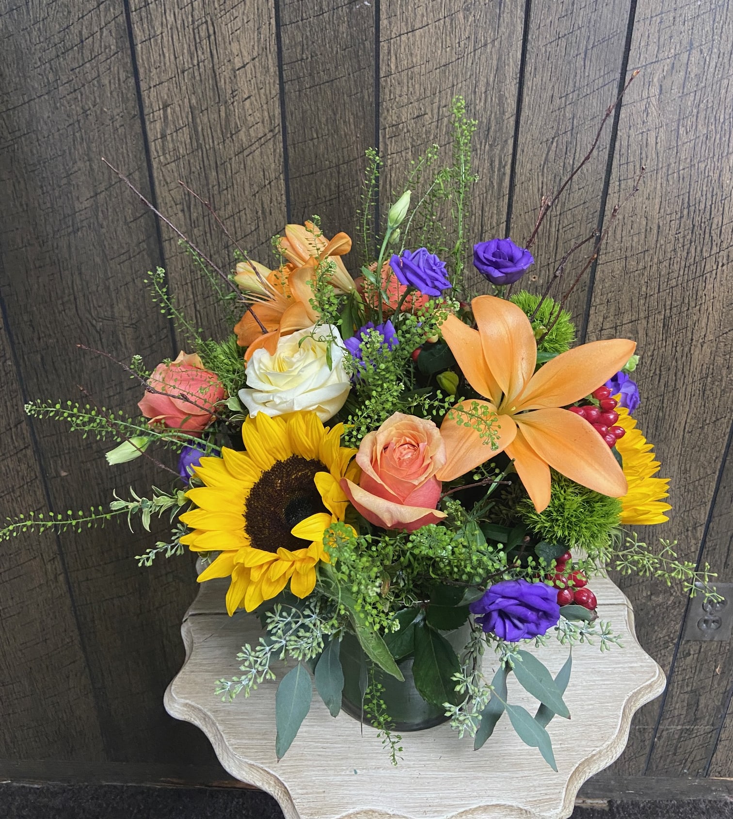 Mixed bouquet with sunflowers, orange lilies, roses, and purple blooms in a glass vase