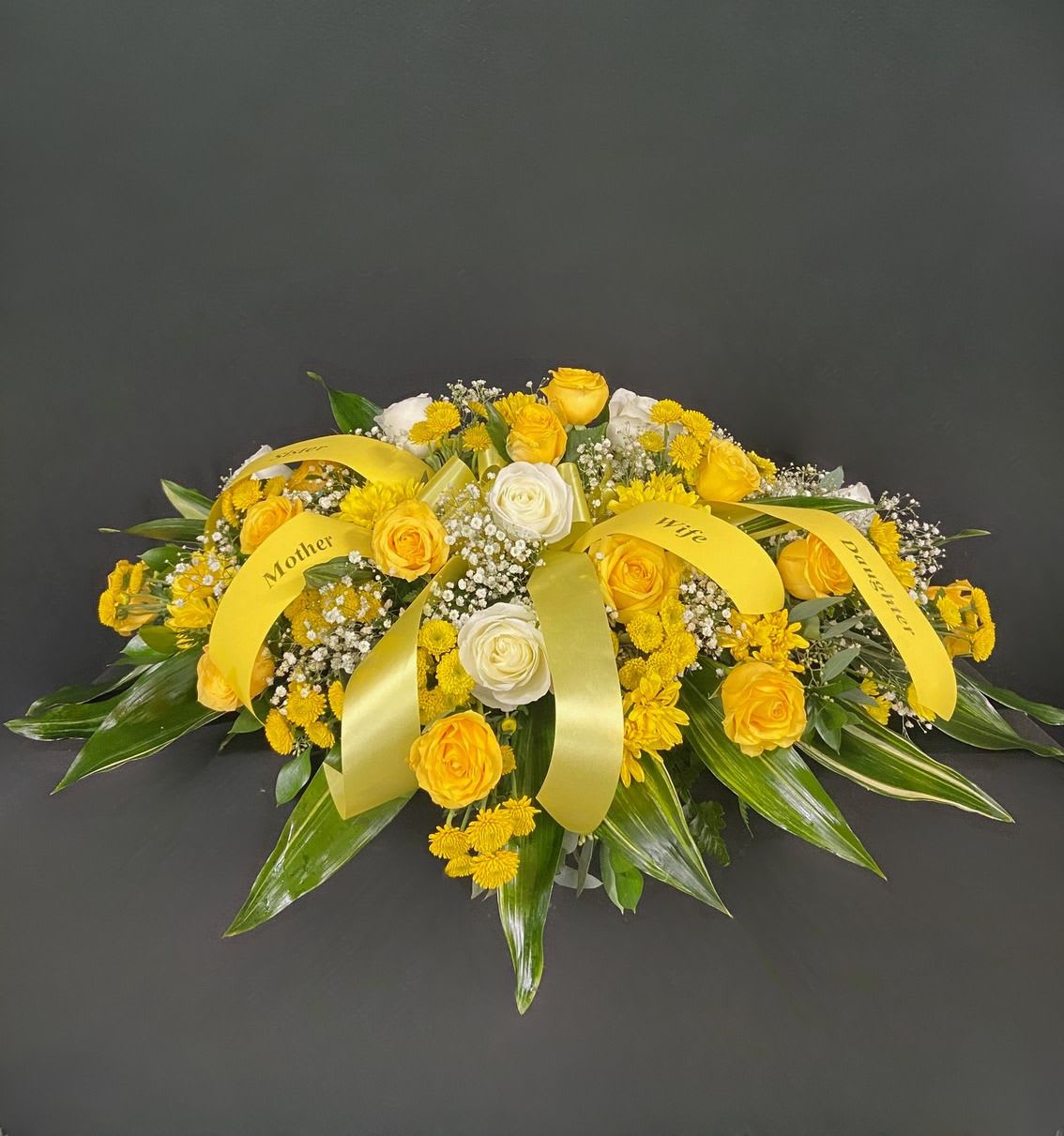 Golden Road Casket Spray - Golden yellow roses become even more striking against a backdrop of elegant white baby's breath accented with button and daisy poms throughout and a ring of white roses across the top. Price includes up to 4 custom printed ribbons.