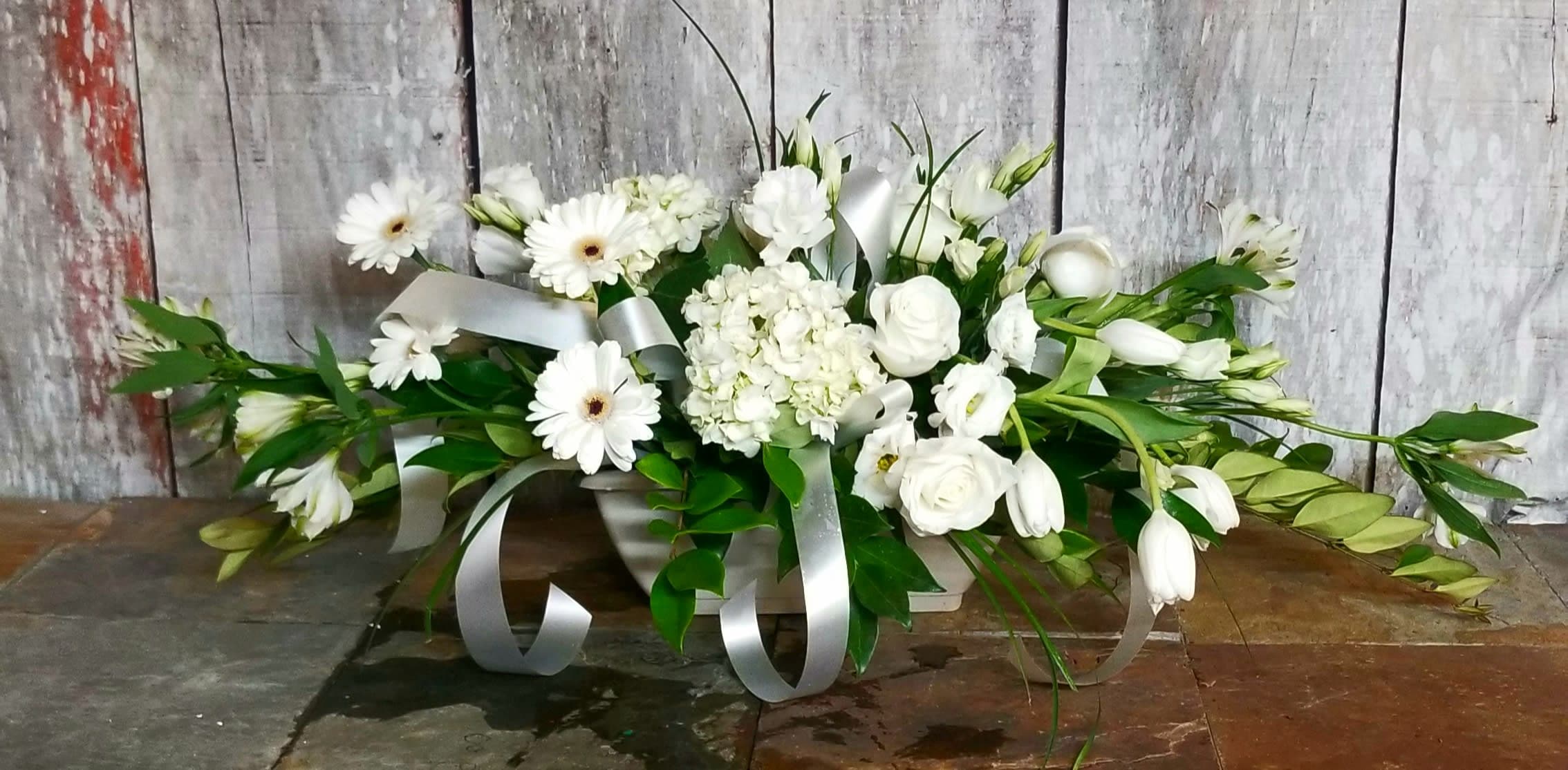 White floral arrangement with daisies, roses, and ribbon in a low container