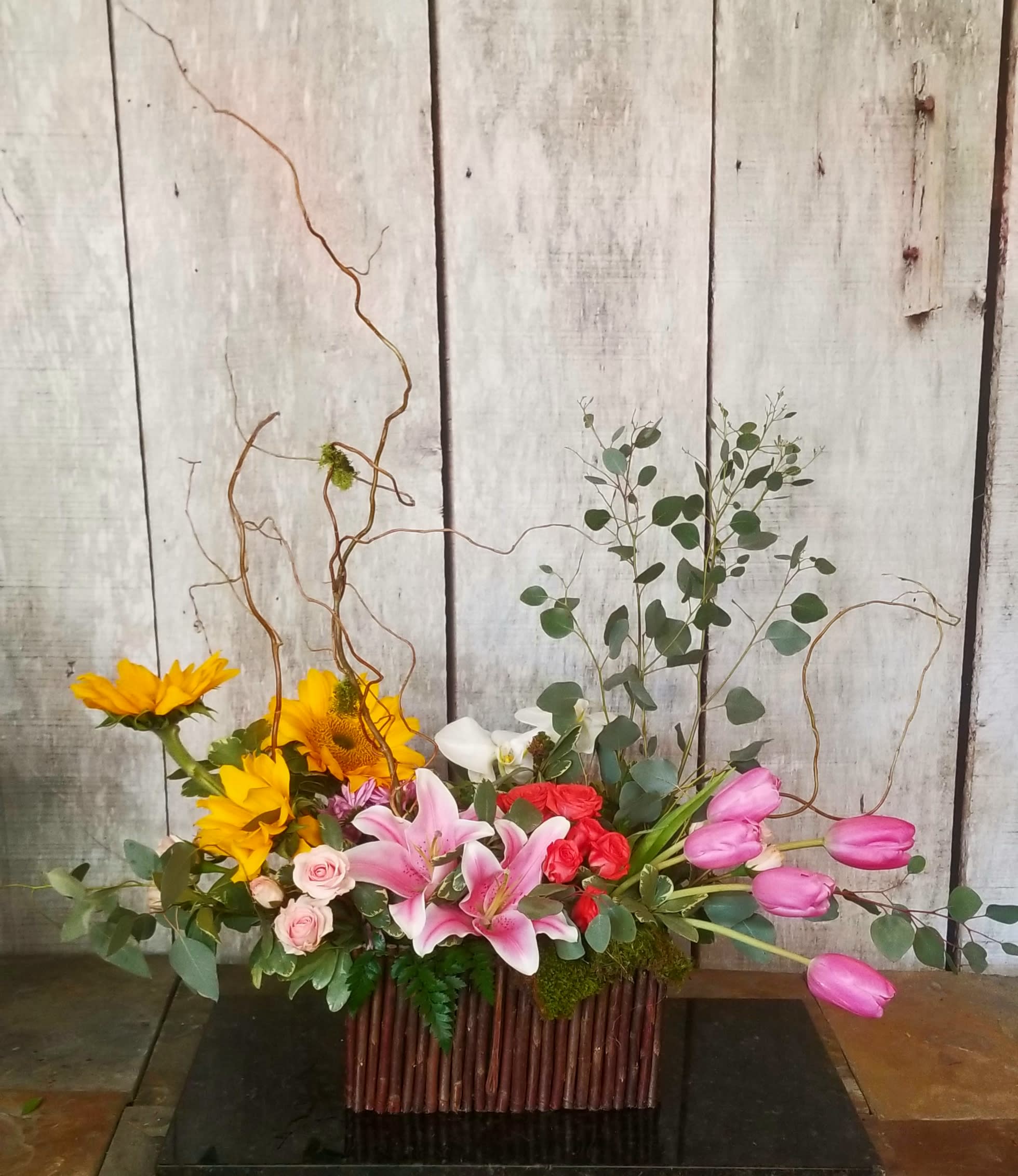 Mixed floral arrangement with sunflowers, lilies, roses, and pink tulips in a rectangular container