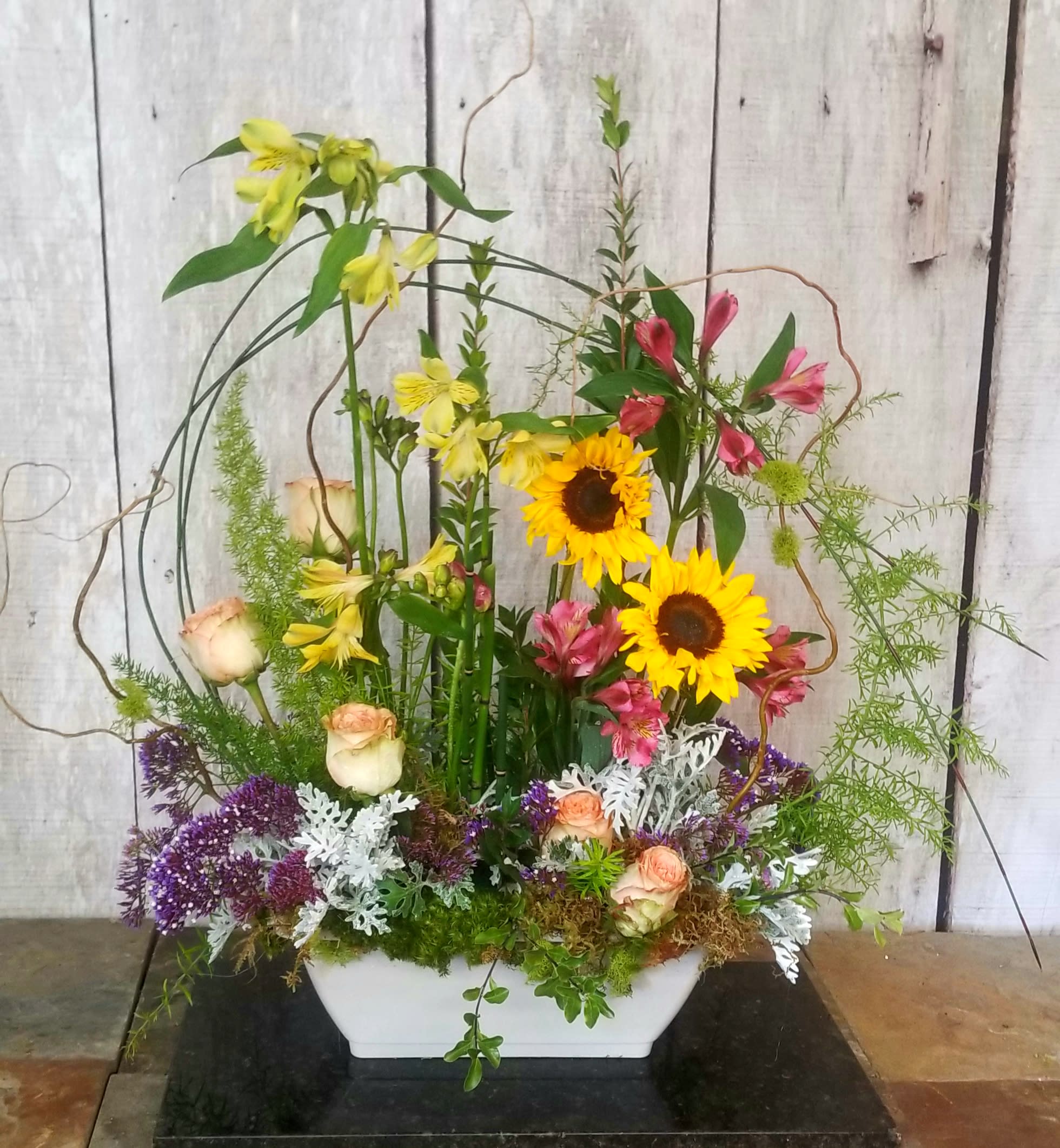 Tall mixed floral arrangement in a white rectangular vase with sunflowers and roses