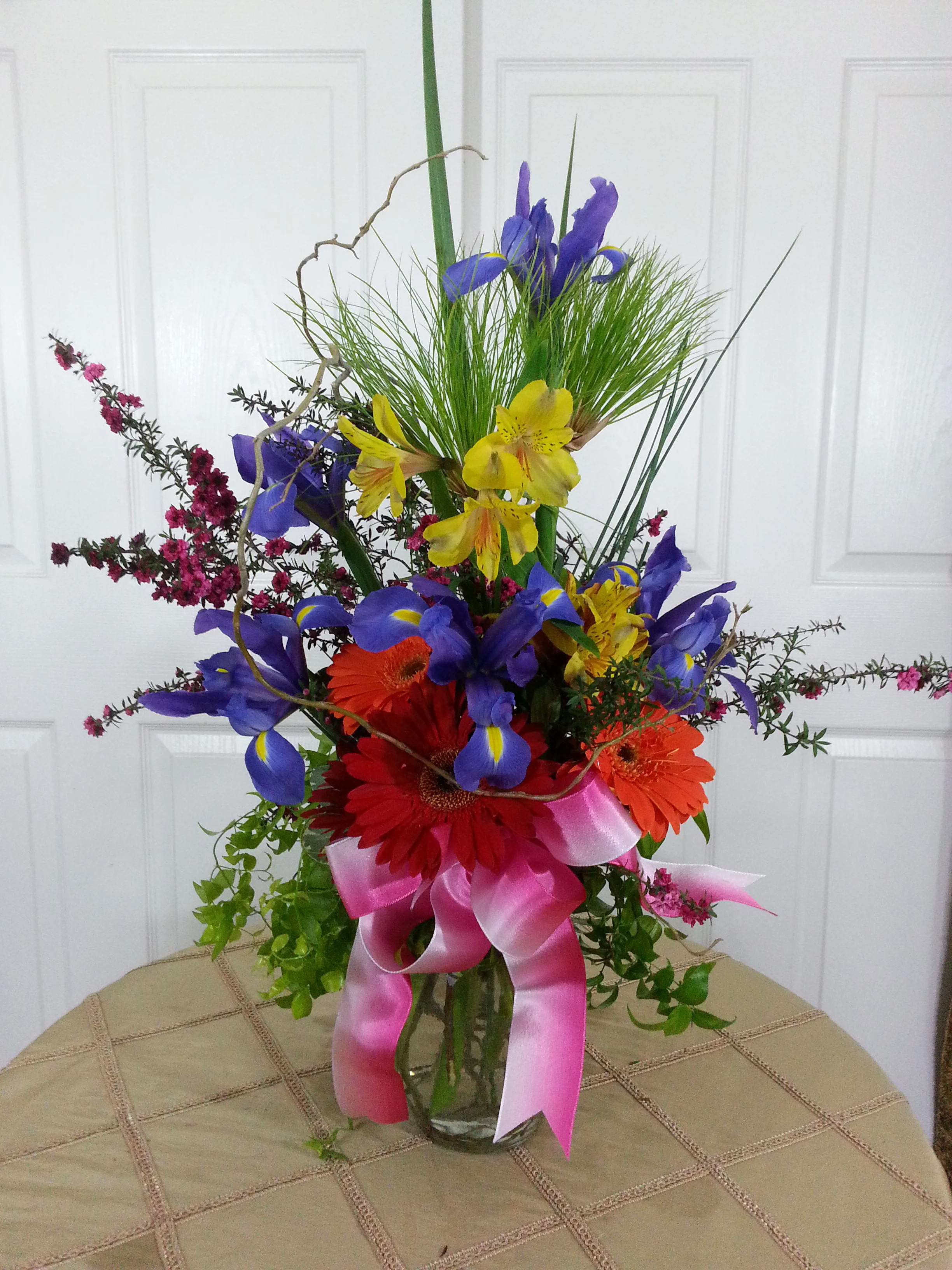 Colorful bouquet with iris, gerbera daisies, and a pink ribbon in a glass vase