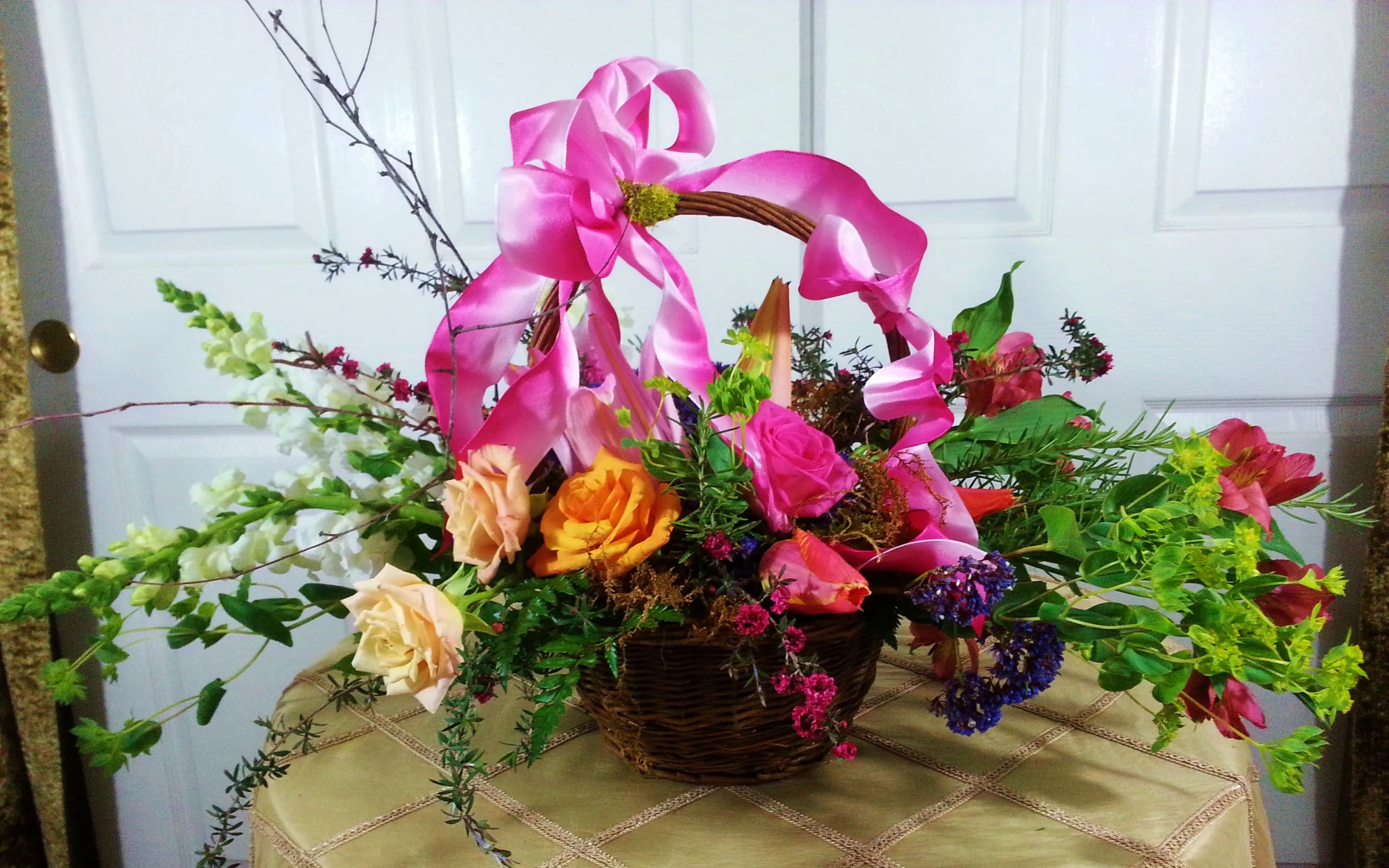 Basket arrangement with roses and mixed flowers topped by a pink ribbon bow