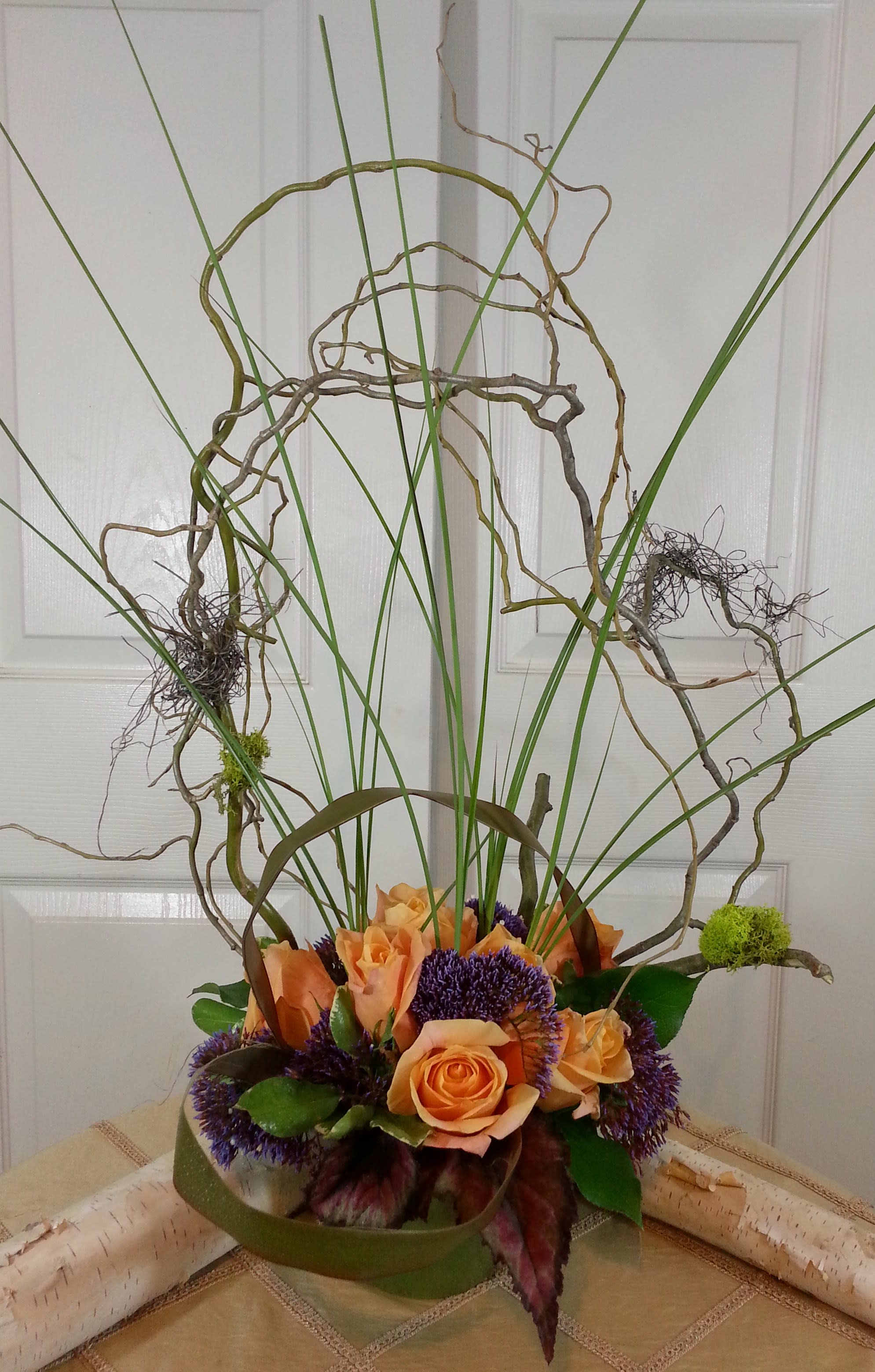 Orange roses with purple blooms in a low green container and tall curly branches
