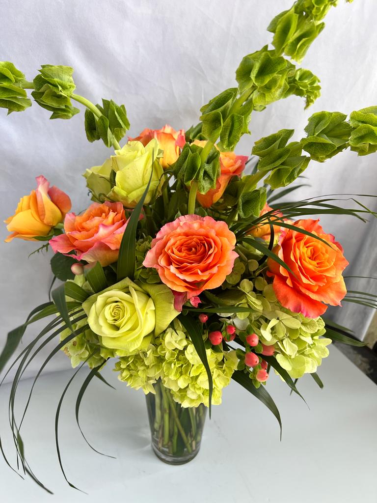 Tall arrangement of orange and green roses, green hydrangeas, and bells of Ireland in a clear glass vase