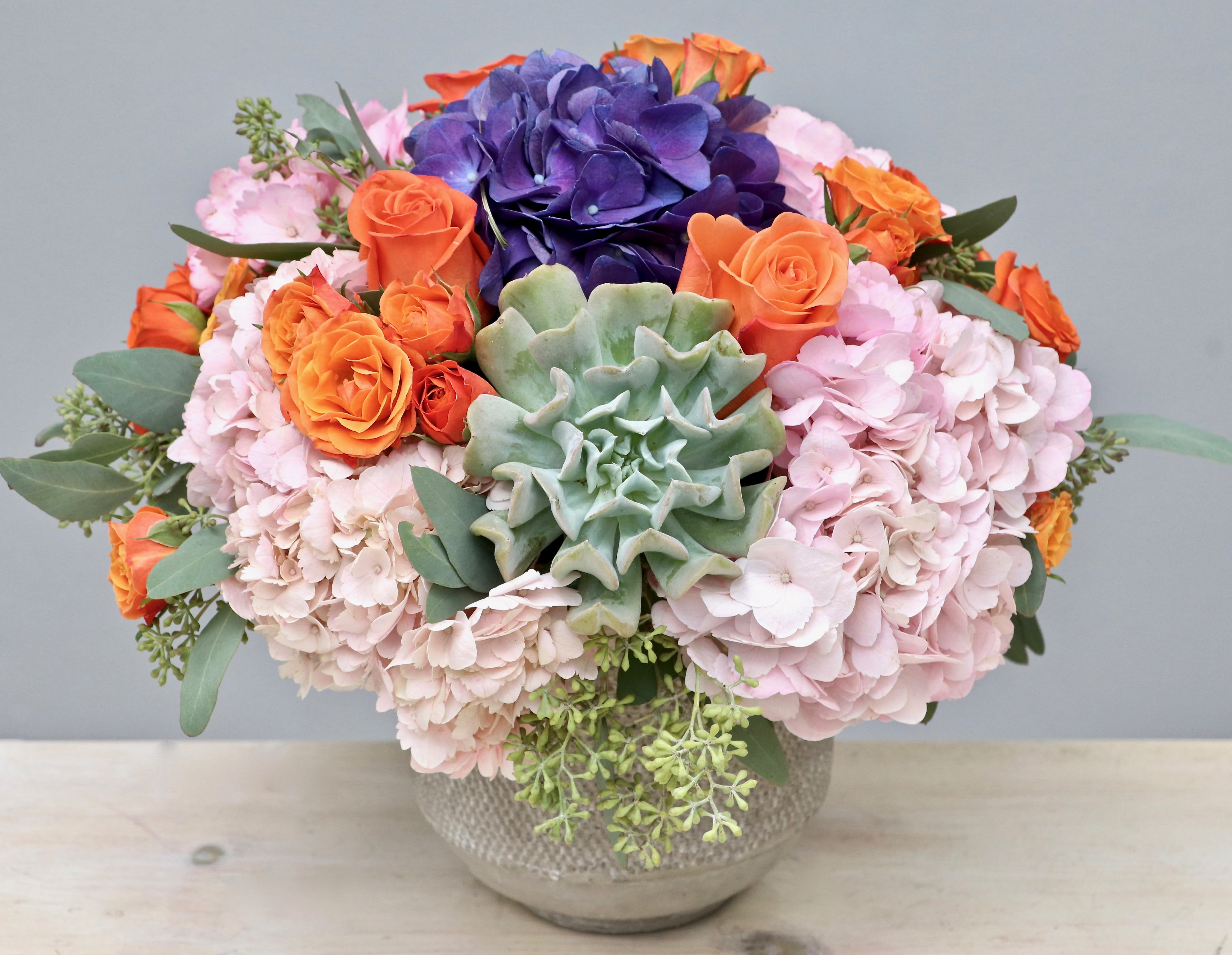 Low bowl arrangement of pink hydrangeas, orange roses, purple blooms, and a central green succulent