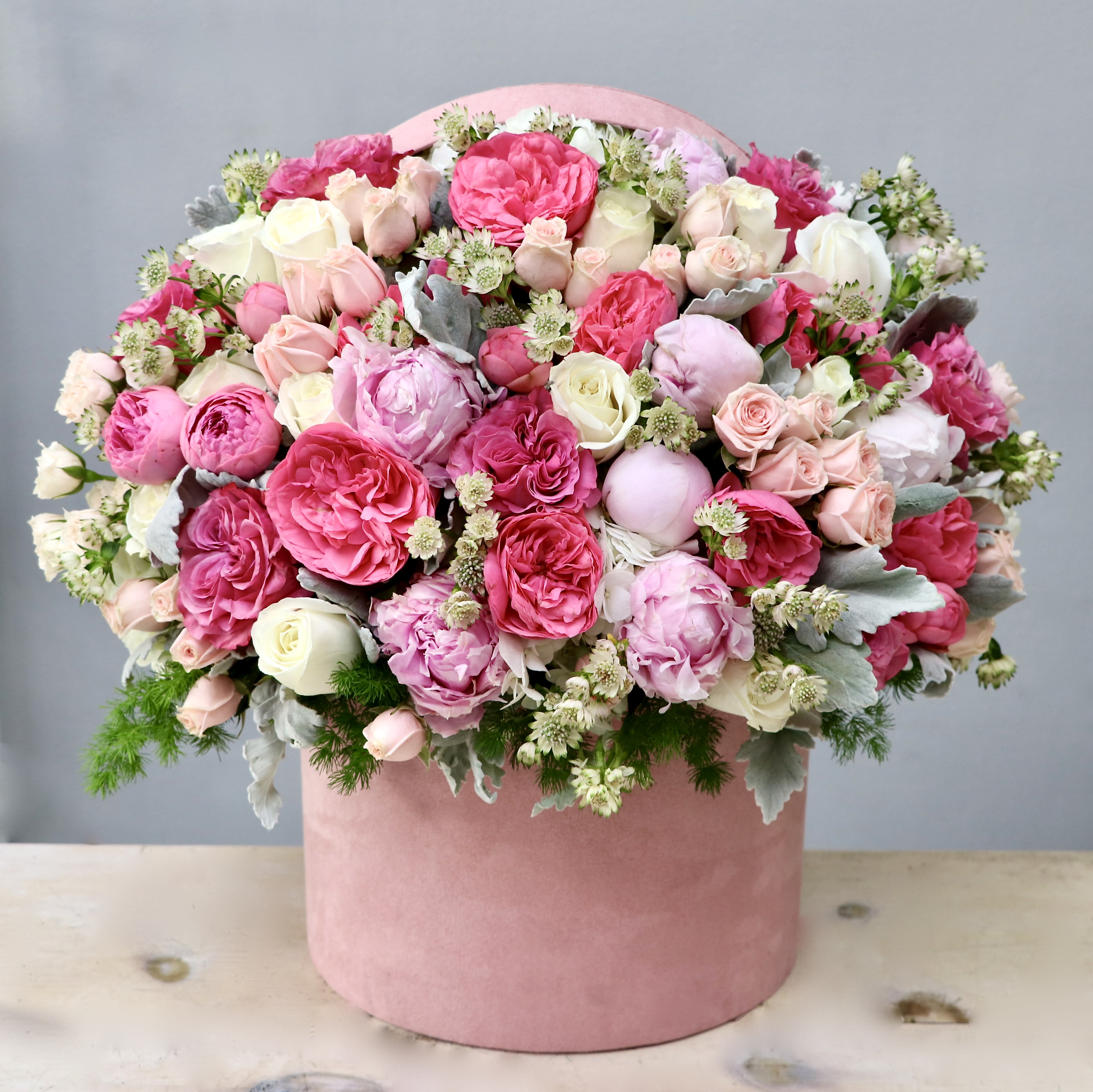 Pink and white roses and peonies arranged densely in a round pink hatbox-style container