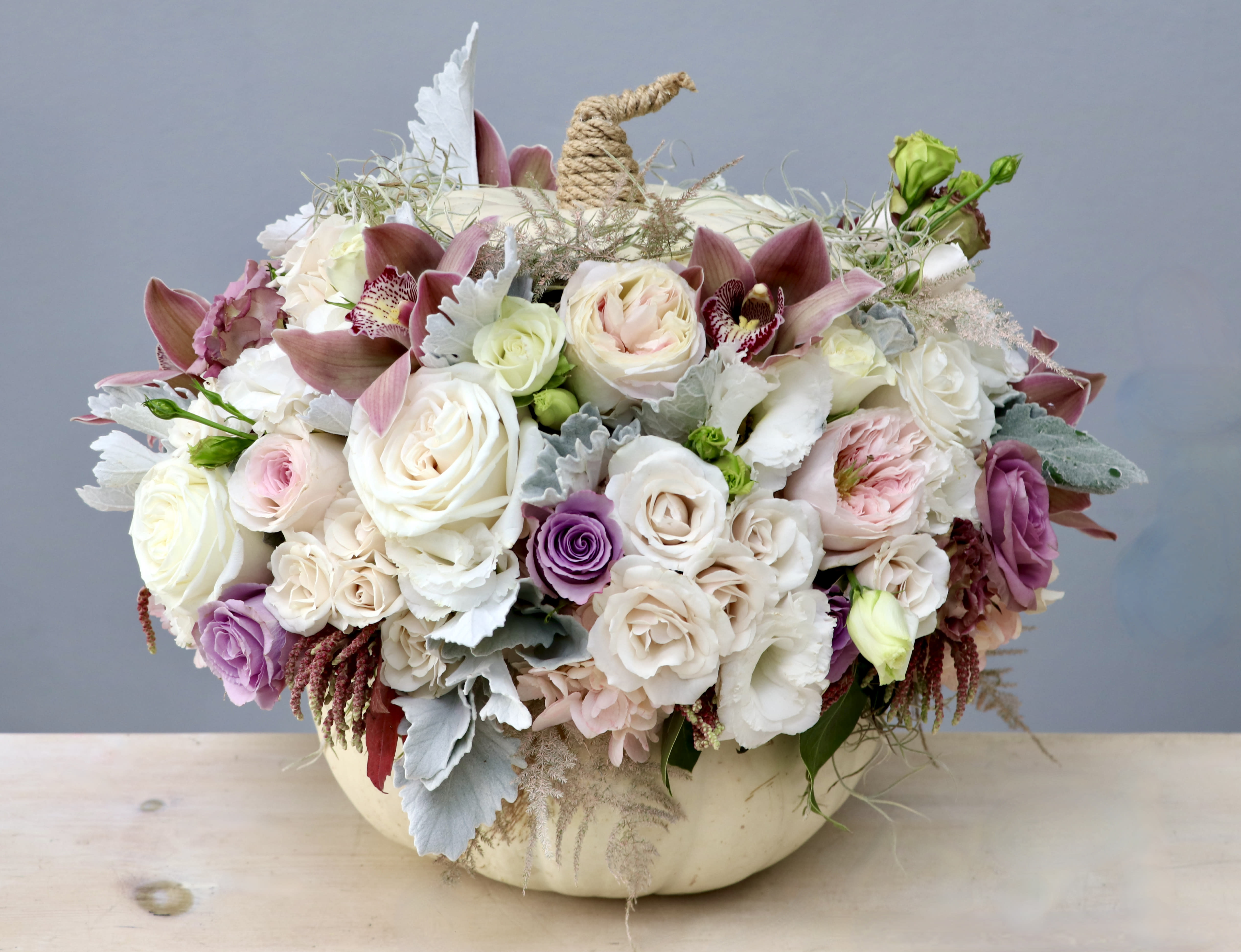 Pastel roses and orchids arranged in a white pumpkin container with a rope stem