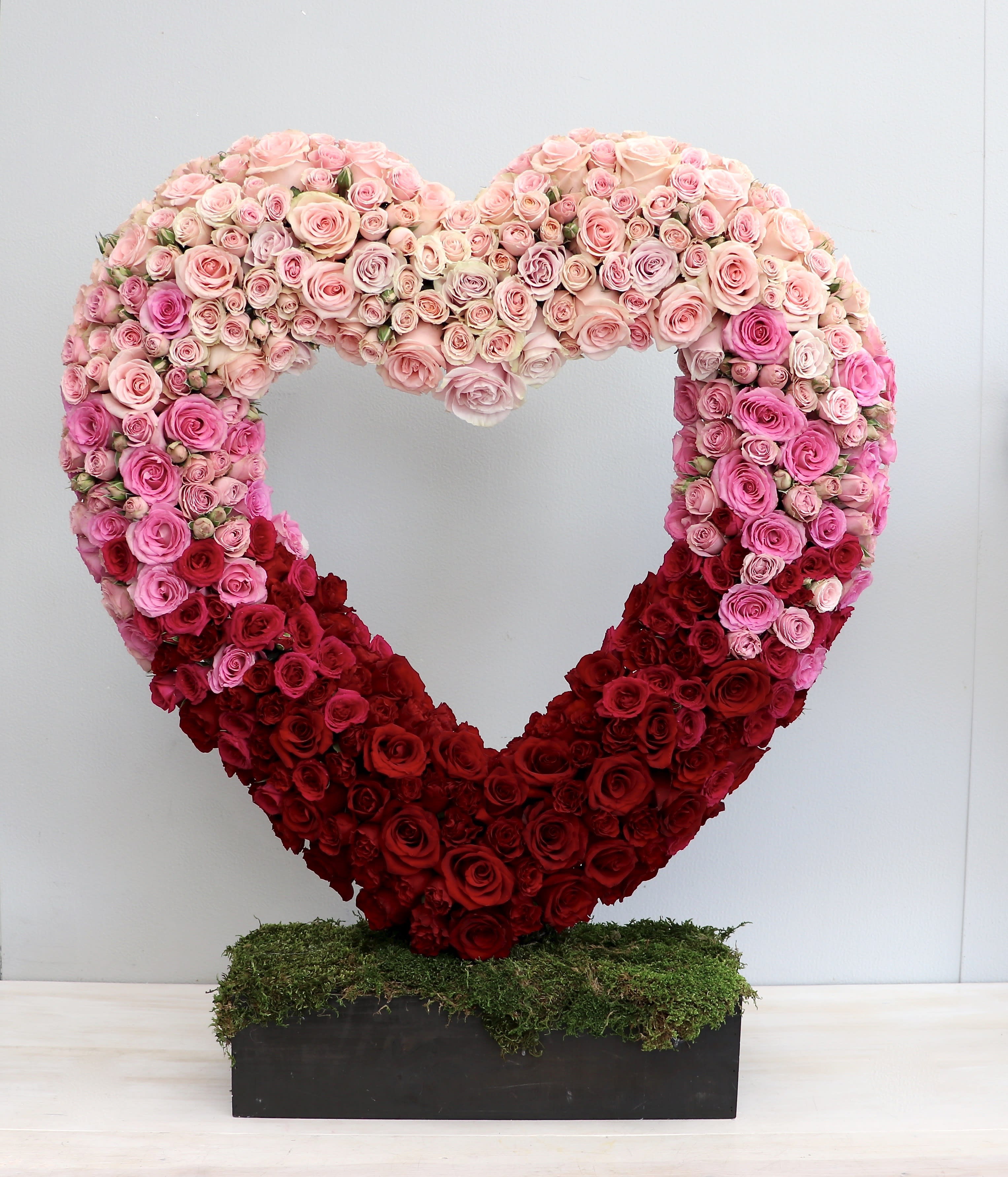Large open heart of pink and red roses on a moss-covered base in a rectangular black container