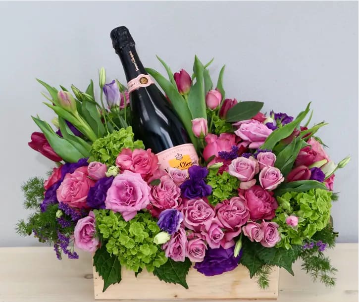 Wooden box arrangement of pink and purple flowers with a champagne bottle nestled in the center
