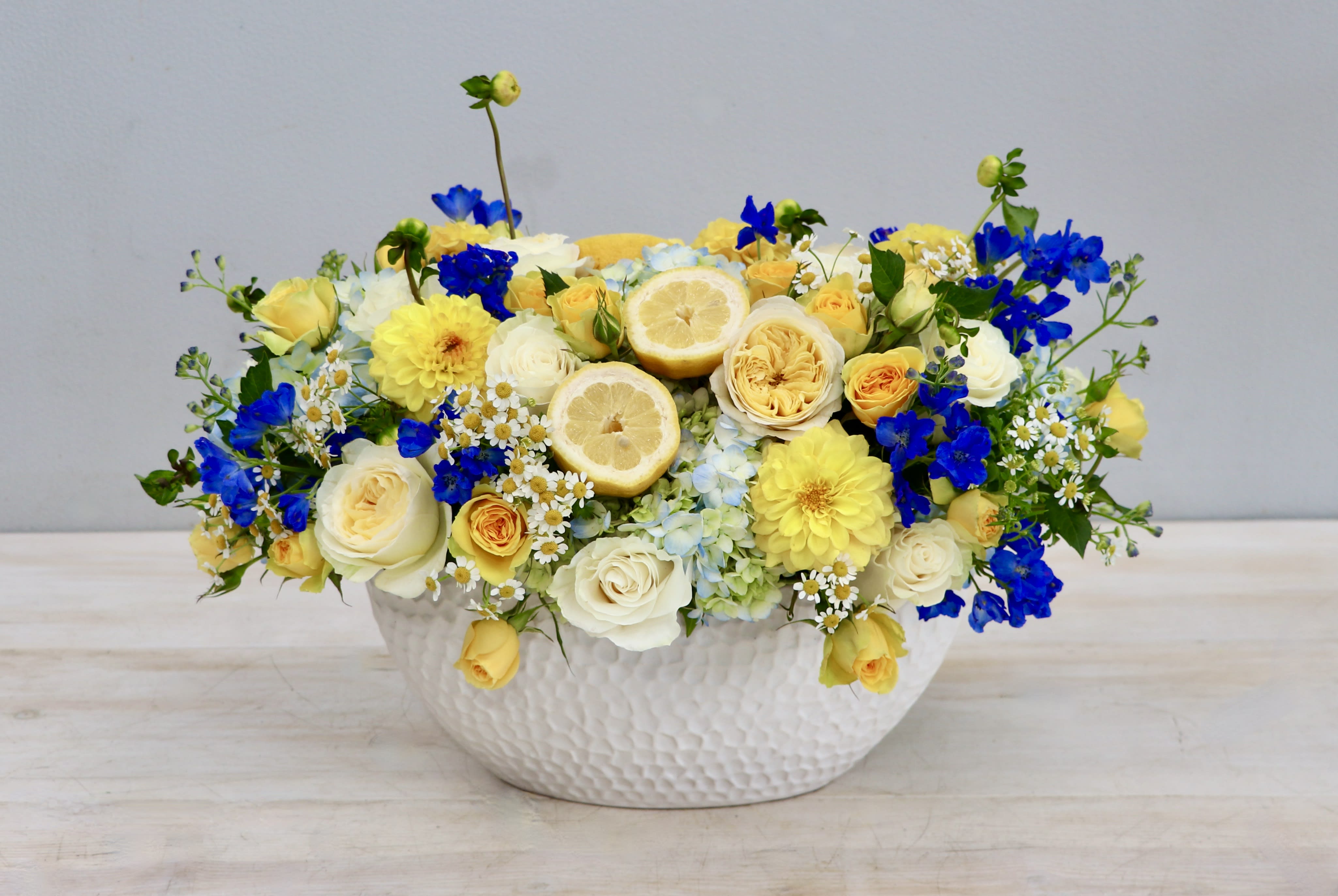 Low yellow, white, and blue flower arrangement with lemon slices in a white ceramic bowl
