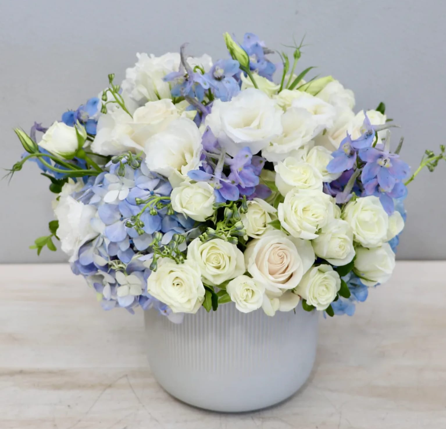 Low white ceramic vase with white roses, blue hydrangeas, and light blue flowers in a rounded arrangement