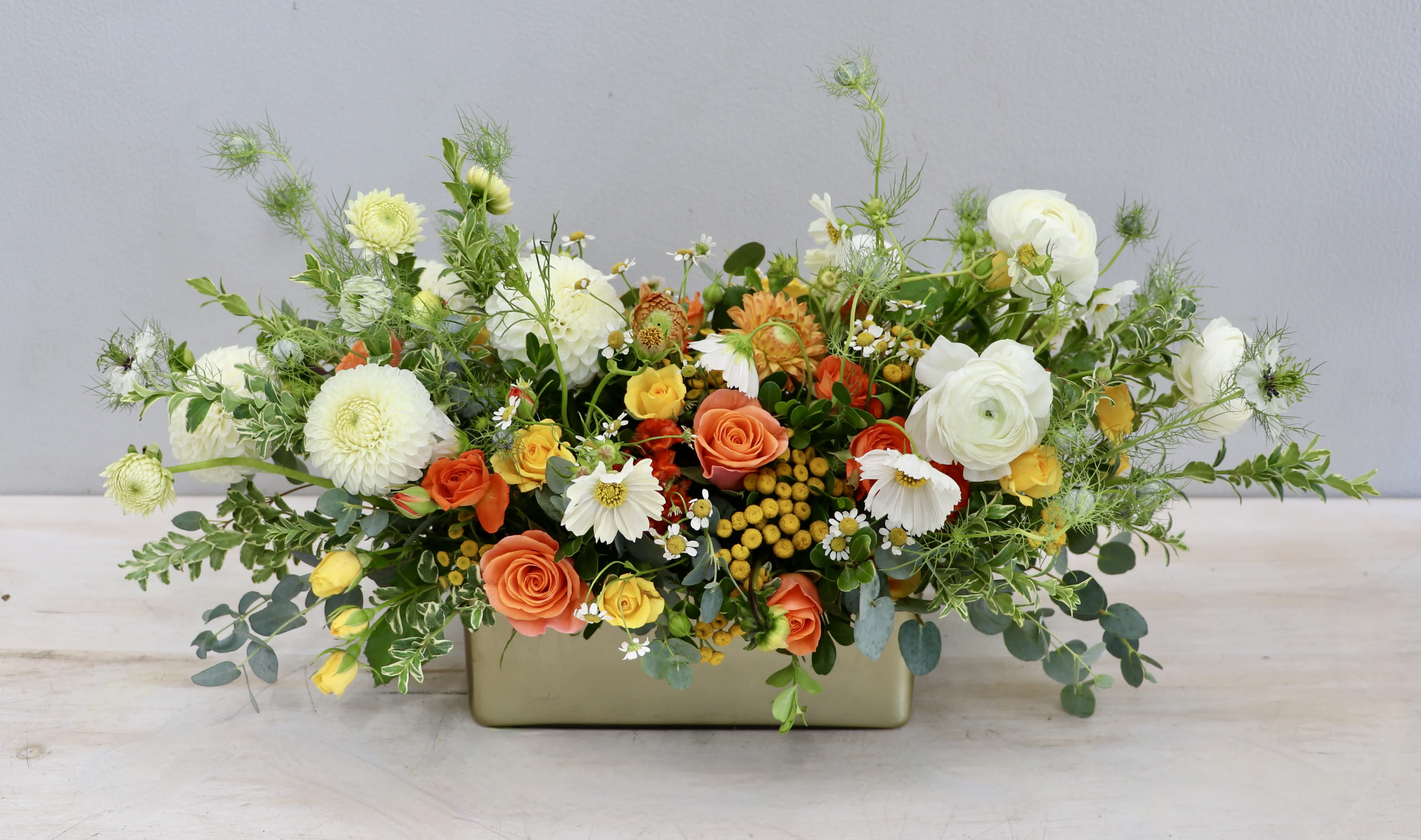 Low centerpiece of white dahlias, orange and yellow roses, and daisies in a gold rectangular vase