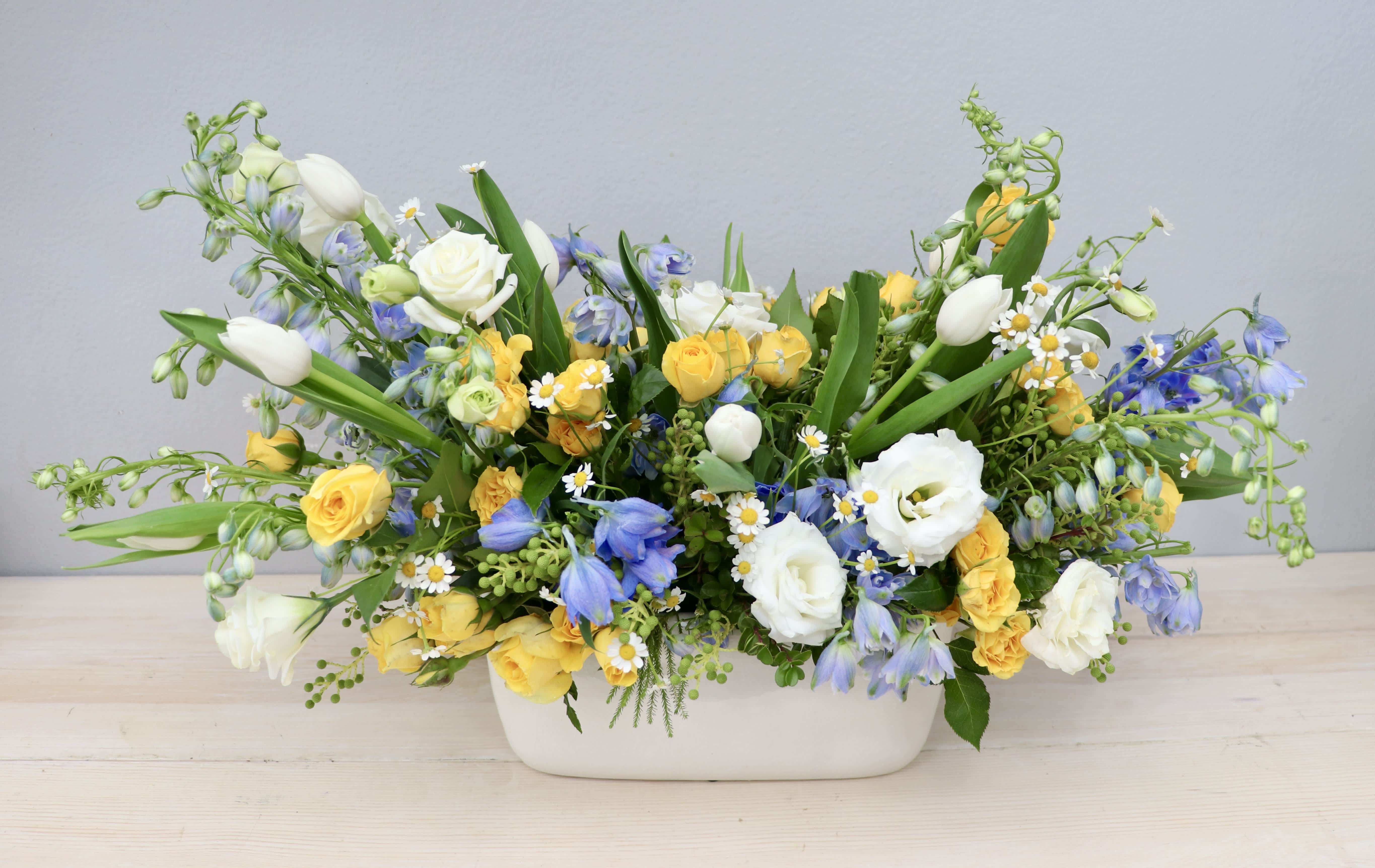 Low centerpiece of yellow roses, white tulips, and blue flowers in a white ceramic container