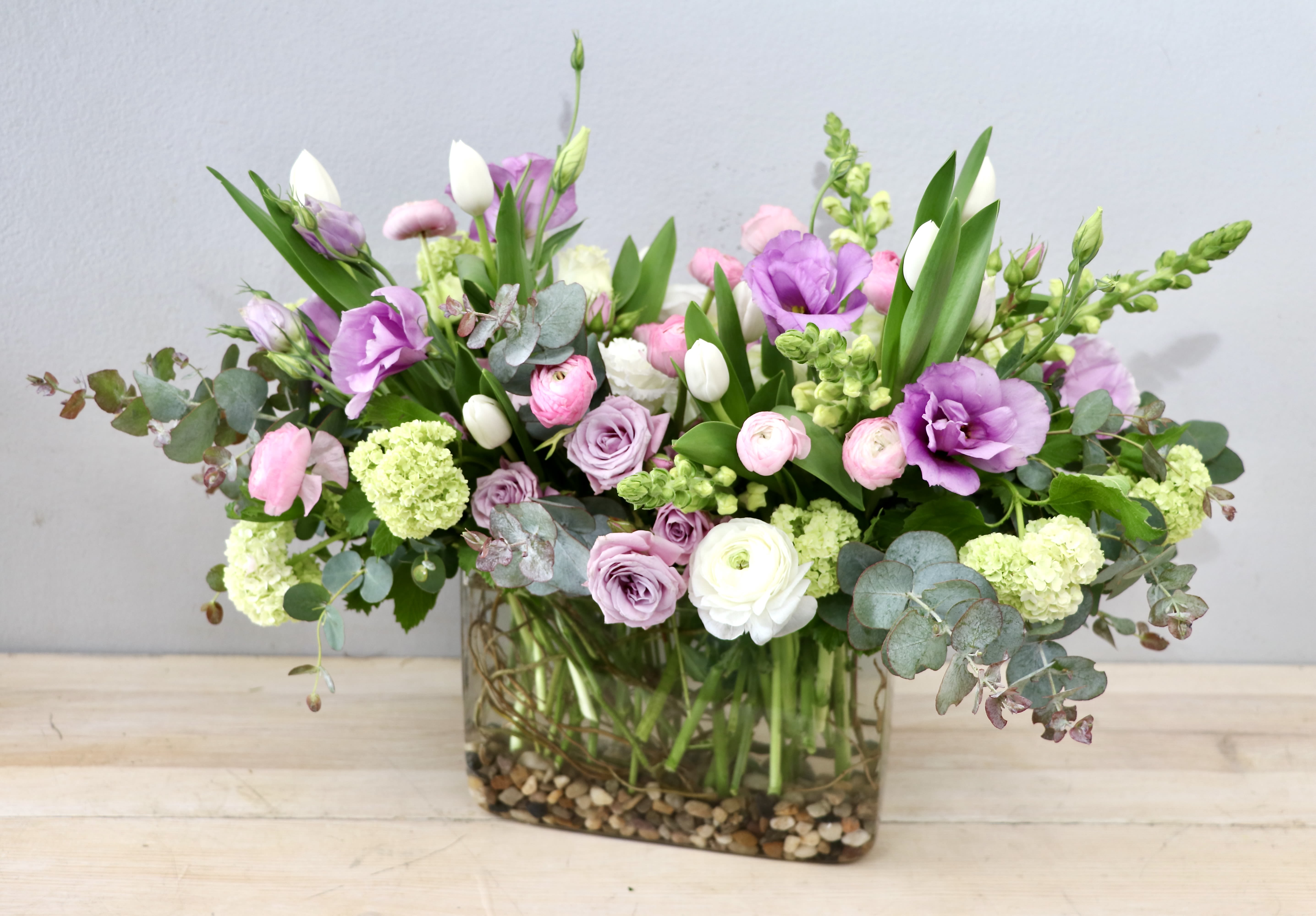 Glendale Meadow - Glendale Flowers - Lavender and blush tulips mingle with ruffled ranunculus and creamy roses in a wide, low garden-style arrangement-perfect as a spring centerpiece or a romantic birthday bouquet. This flower arrangement for everyday gifting or celebratory occasions places those soft pinks, purples, whites, and fresh greens right up front so the person you're thinking of will feel seen and cherished the moment it arrives.  The mix includes tulips, ranunculus, roses, lisianthus, and snapdragons arranged with pale eucalyptus-like foliage for airy texture and gentle movement. Blooms are gathered in a clear rectangular glass vase with decorative pebbles visible at the base, creating a natural, grounded look that suits tabletops and mantels.  Made to sit wide, this composition works beautifully as a dining or reception centerpiece or a thoughtful gift to mark a birthday, anniversary, or just-because moment. For best results, place in a cool spot away from direct heat and refresh the water every couple of days to keep blooms looking fresh.