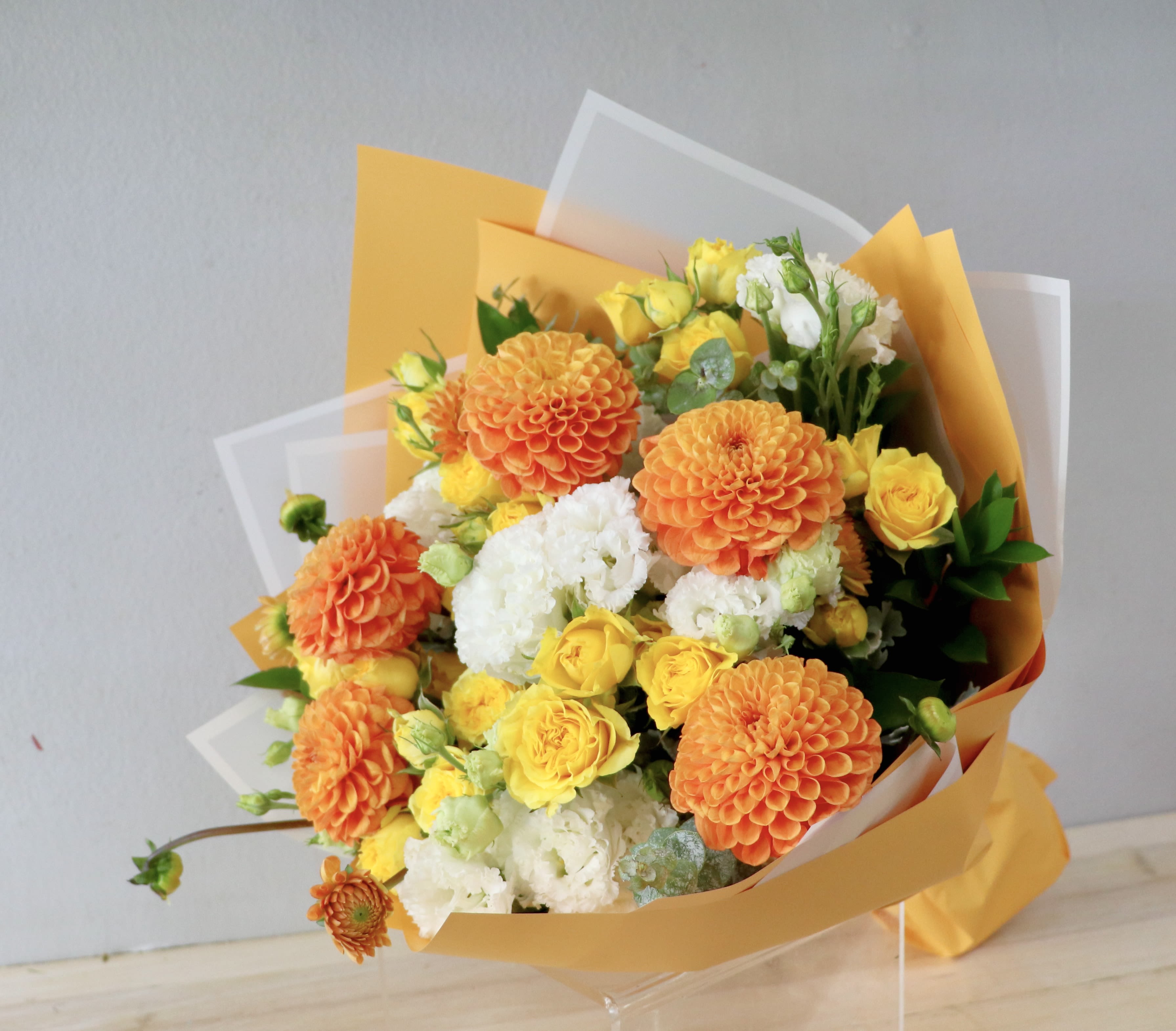 Apricot Sunset - Glendale Flowers - Brighten any space with this vibrant, garden-style bouquet. Featuring striking orange pompon dahlias, cheerful yellow spray roses, and crisp white lisianthus, this arrangement captures the warmth of a summer afternoon. Accented with fresh eucalyptus and nestled in layers of golden-orange and frosted wrapping, it offers a lush, textured look perfect for birthdays, celebrations, or a joyful everyday surprise.