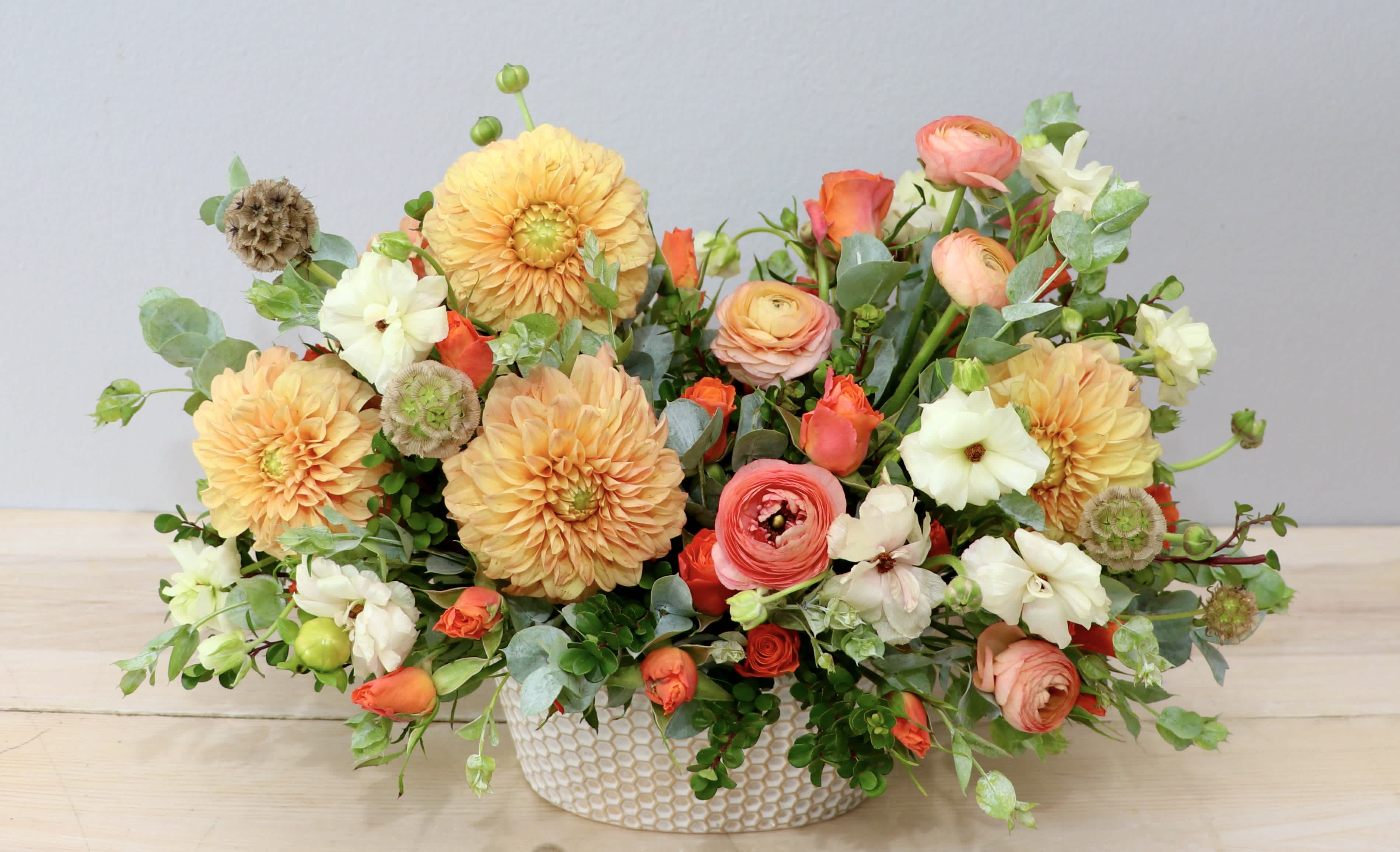 Golden Hour - Glendale Flowers - Featuring focal blooms of golden-peach dahlias and layer upon layer of coral ranunculus, this low-profile centerpiece is accented with delicate white butterfly ranunculus, spray roses, scabiosa seed pods, and silvery eucalyptus foliage.