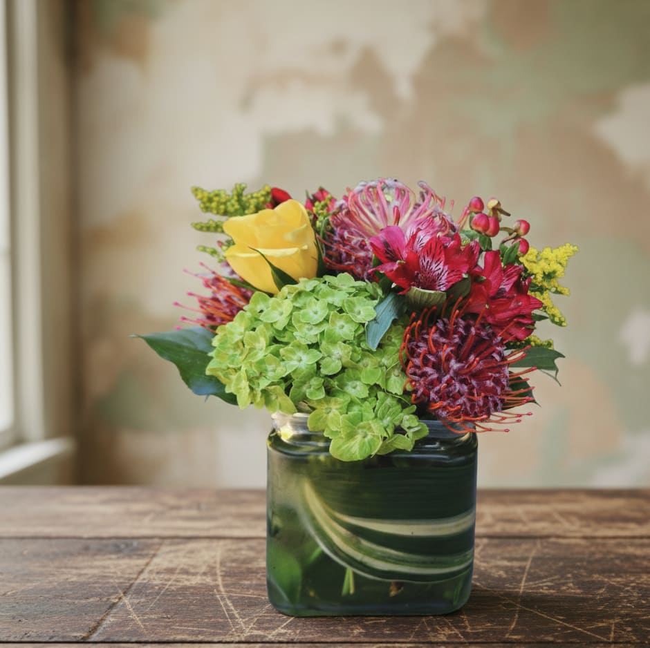 Compact arrangement of green hydrangeas, yellow calla, and red protea in a square glass vase