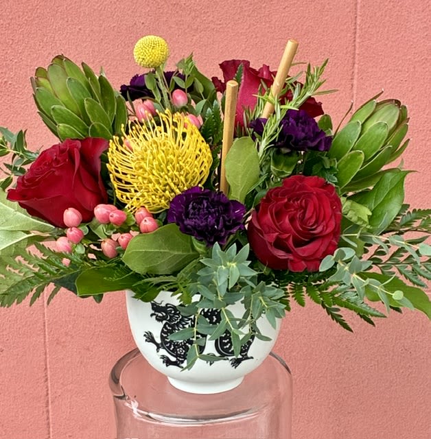 Year of the Dragon Pho Bowl - Bold, sculptural blooms make an unforgettable birthday or anniversary gift that reads as joyful and confident. This flower arrangement centers on two prominent red roses and a bright yellow pincushion protea, accented with deep purple blooms and small pink berry-like fillers to create a layered, garden-style composition that says "celebrate" without being fussy. The overall effect is vivid and cheerful-perfect for an anniversary, a birthday, or a "just because" surprise that should make the person you're thinking of feel truly seen.  Handcrafted in a white ceramic vase with a black decorative motif, the design is framed by broad green foliage for a lush, textural look that stands tall on a dining table or entry console. Protea and roses provide striking form and longevity while the mix of colors-red, green, purple, and yellow-keeps the arrangement lively and versatile. Arrives ready to display; simple care (fresh water and a cool spot away from direct sun) will help the blooms last so your recipient can enjoy the rich colors for days after the occasion.