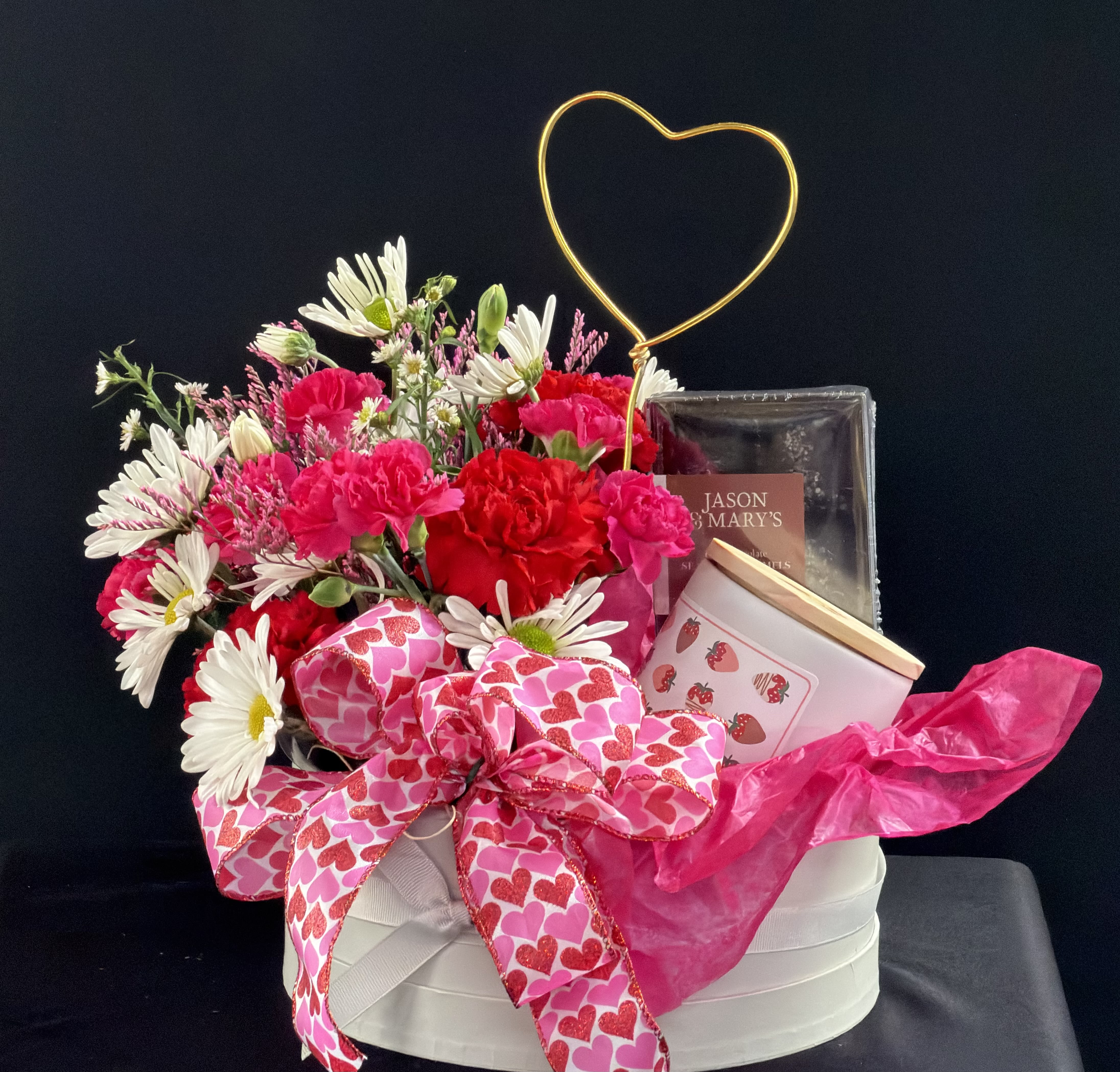 Pink and white flower arrangement with a heart topper and gift items in a round box