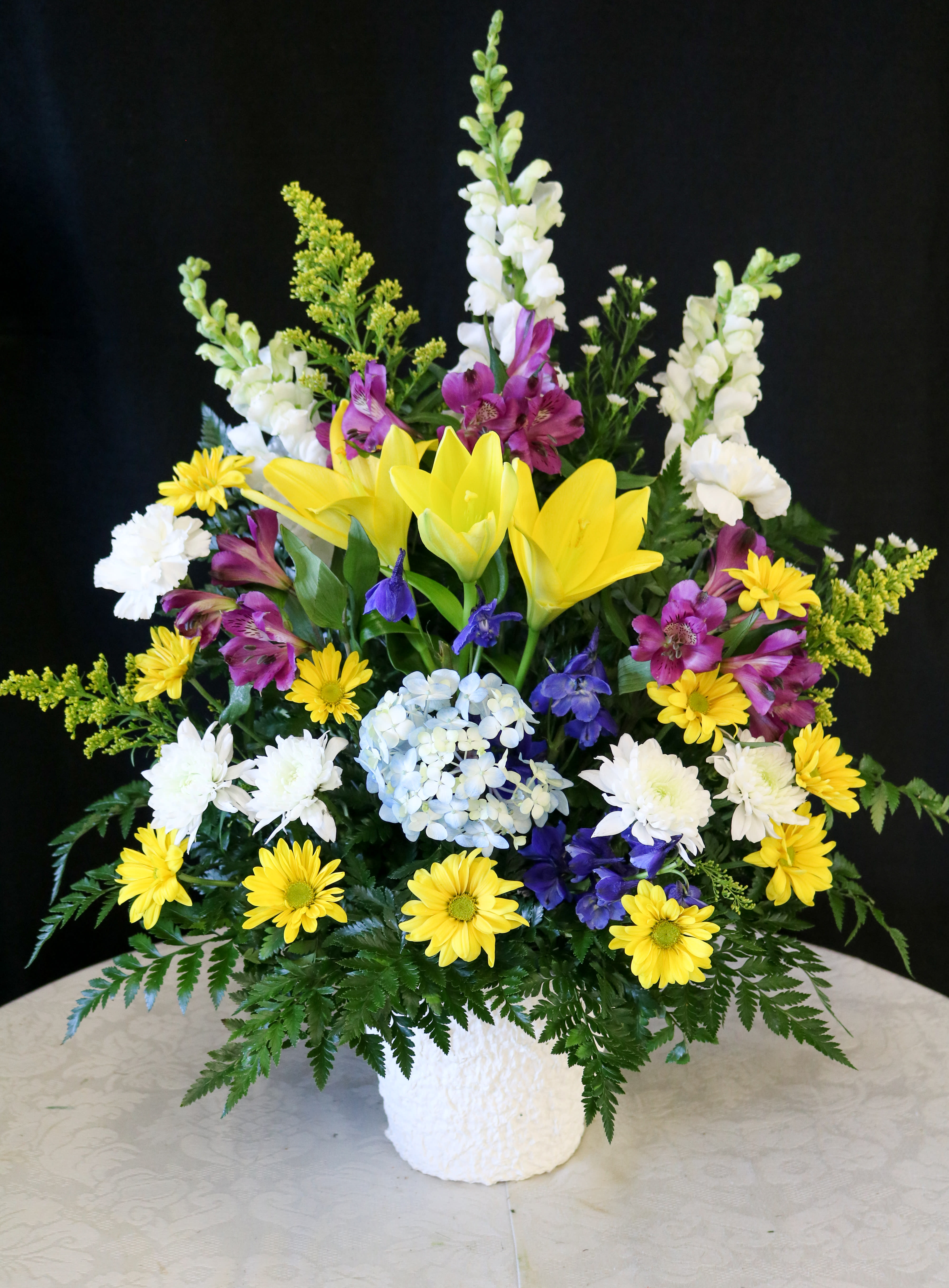 Mixed floral arrangement with yellow lilies, white blooms, and purple flowers in a white vase