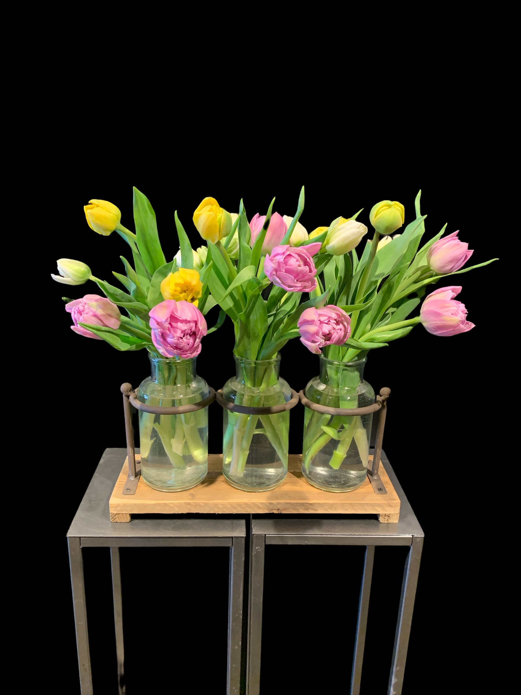 Pink and yellow tulips arranged in three clear glass bottle vases on a wood and metal stand