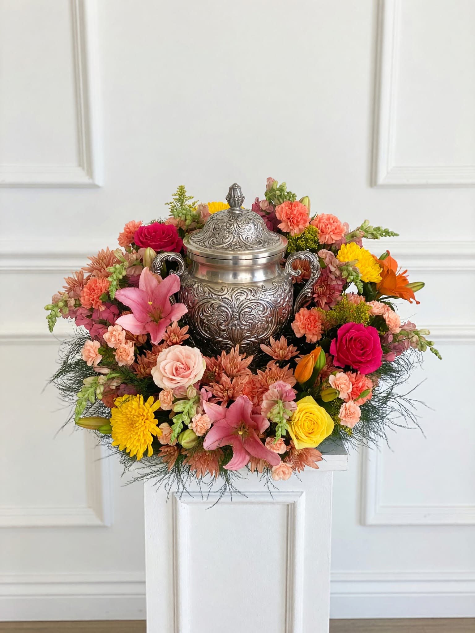 Vibrant Urn Arrangement  - Roses, lilies, carnations, chrysanthemums and snapdragons are layered to create a lively, textured mix: soft rose petals, trumpet-shaped lilies, ruffled carnations and pompom chrysanthemums combine with taller snapdragons for subtle vertical movement. The color palette-peach, coral, warm yellow and vivid pink-reads bright and joyful from every angle, with a symmetrical, densely packed composition that looks abundant in large event spaces and photos.