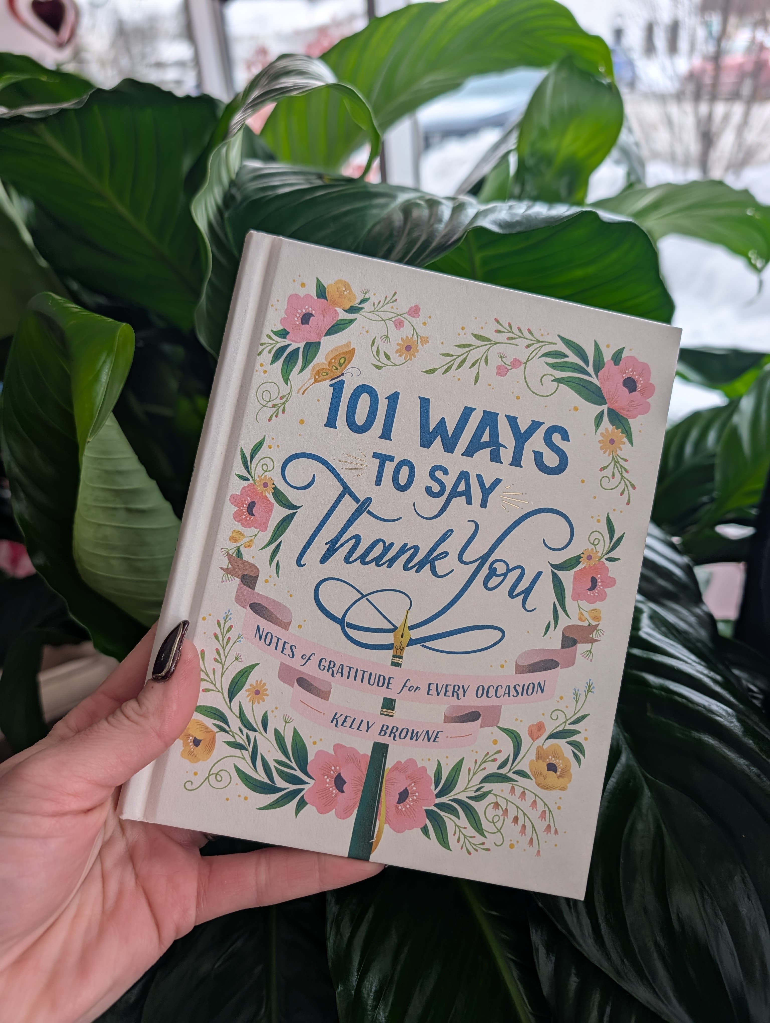 A hand holds a floral gratitude book in front of large green leaves.