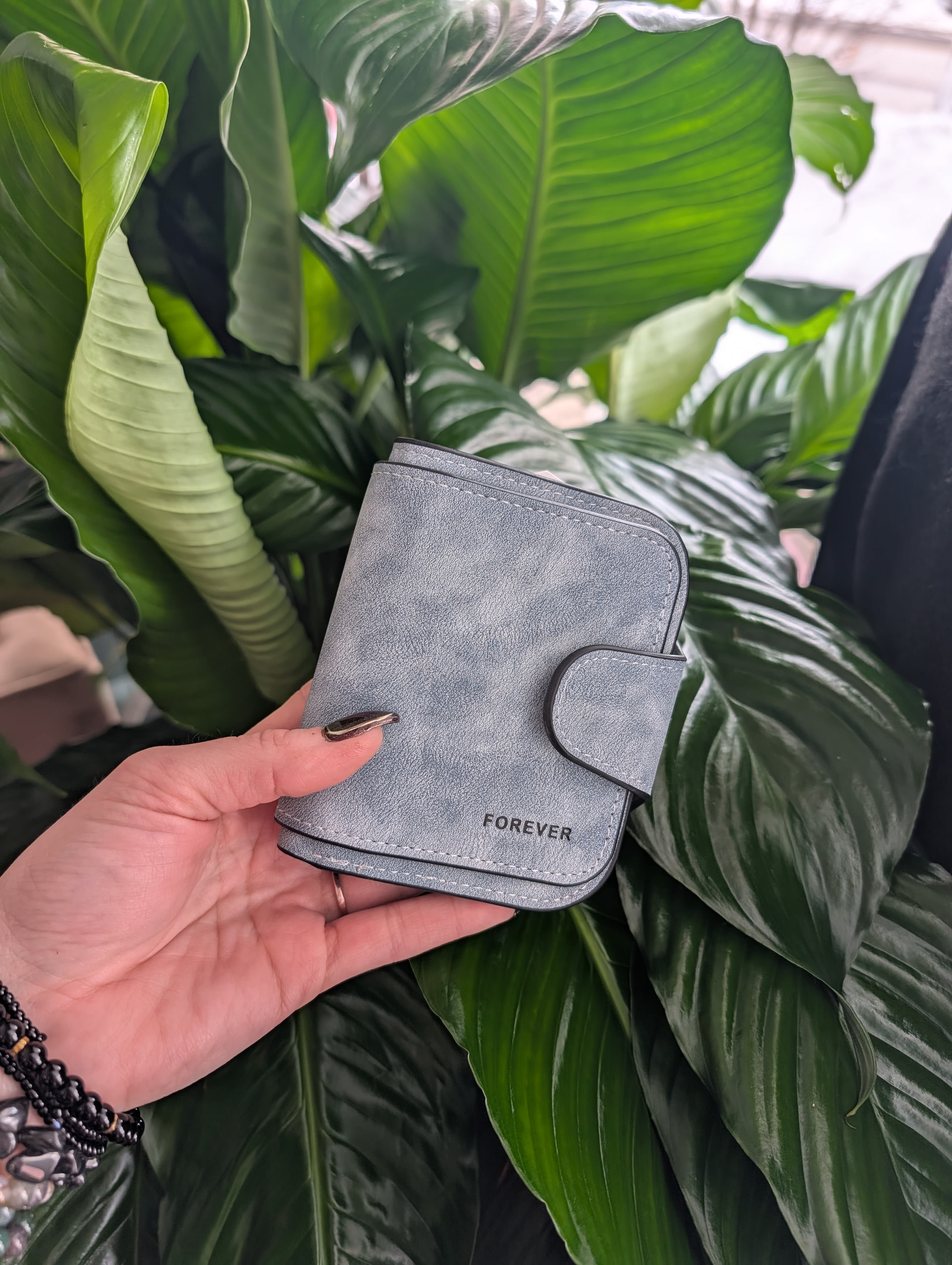Hand holding a gray wallet in front of large green plant leaves.
