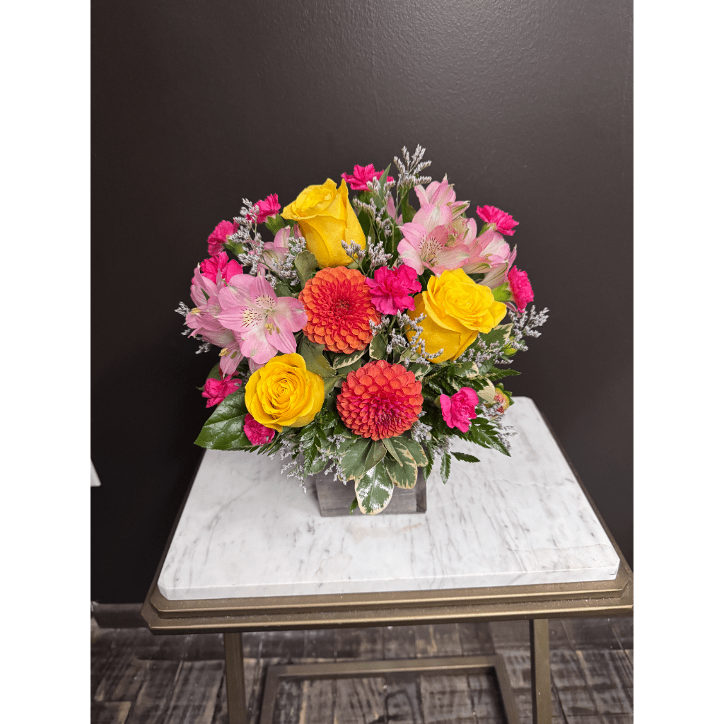 Bright mixed bouquet with yellow roses, pink flowers, and orange blooms in a square vase