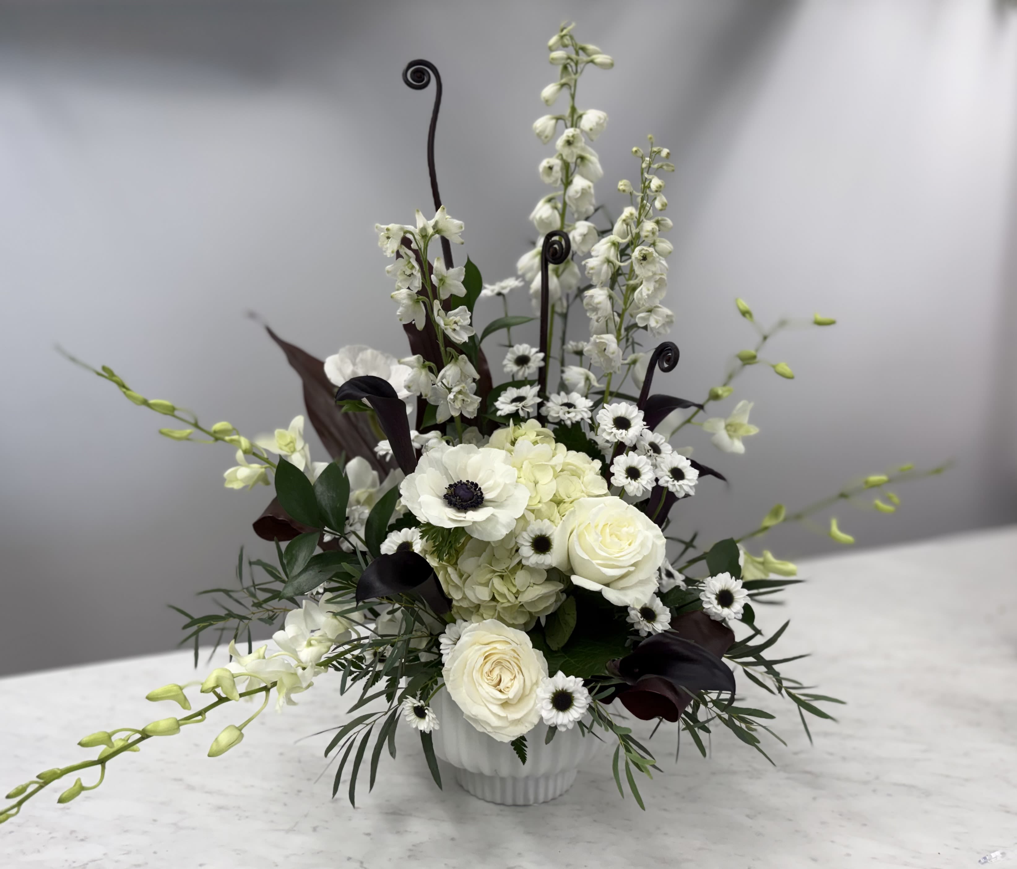 Spooky Compote - “I have no idea what I’m doing, but I know I’m doing it fabulously.” - Moira Rose, Schitt's Creek  This fashion-forward arrangement features white delphinium, dendrobium orchids, uluhe ferns, anemones, black calla lilies, red tai leaves, yin-yang daisies and of course, stunning white Roses front and center.  