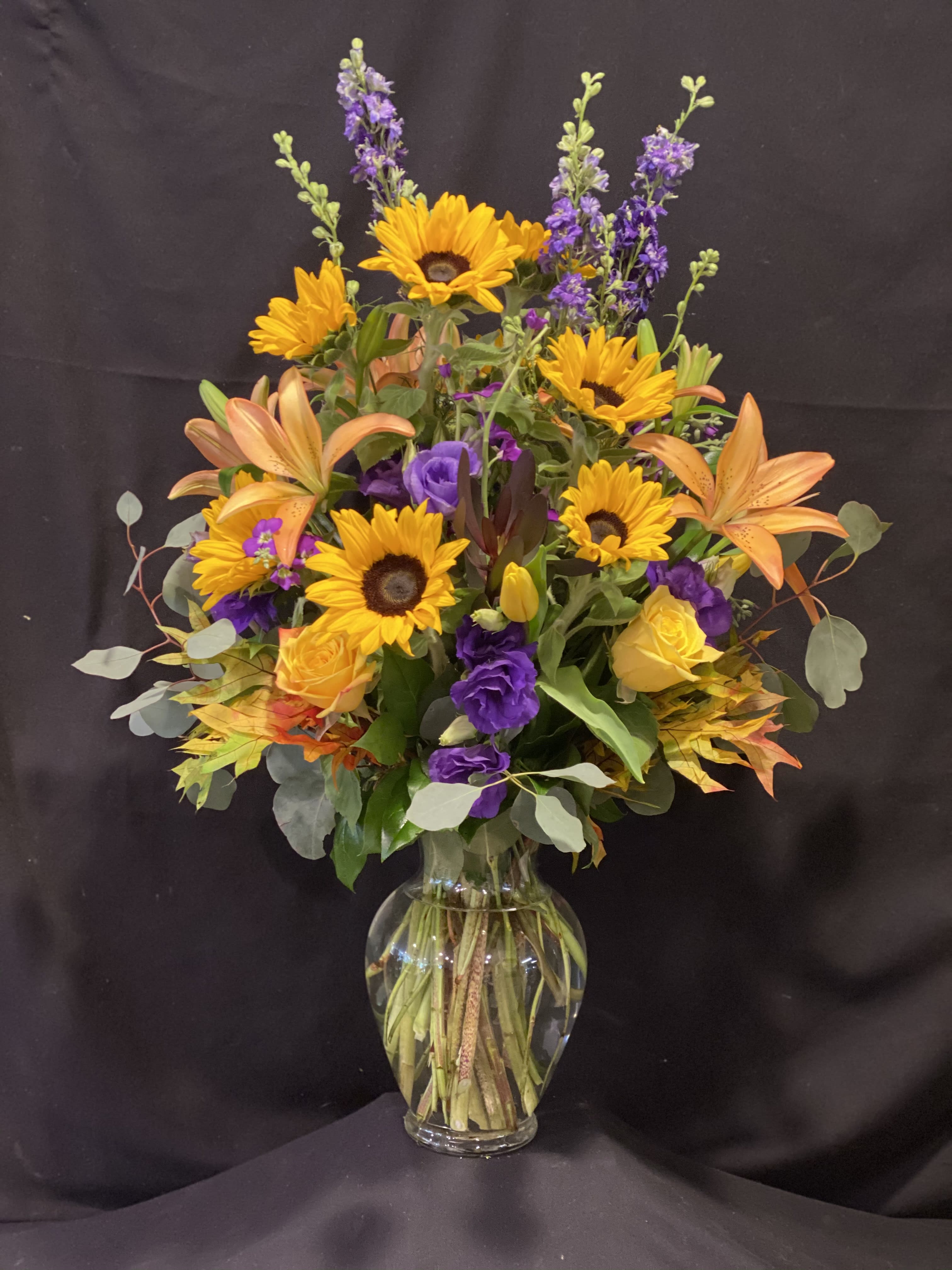 Mixed bouquet of sunflowers, lilies, and purple blooms in a clear glass vase