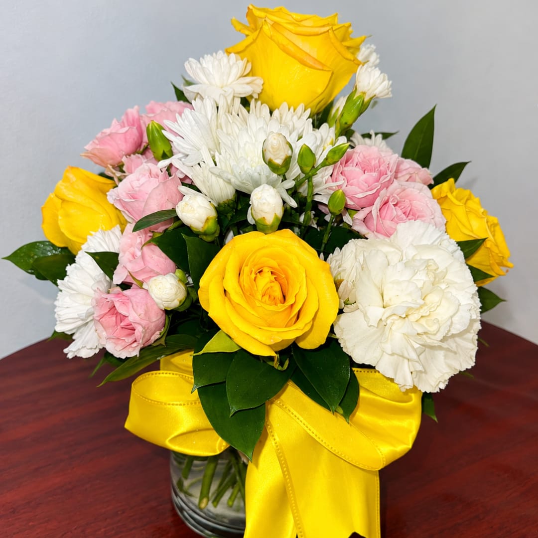 Yellow and pink roses with white daisies in a glass vase with a yellow ribbon