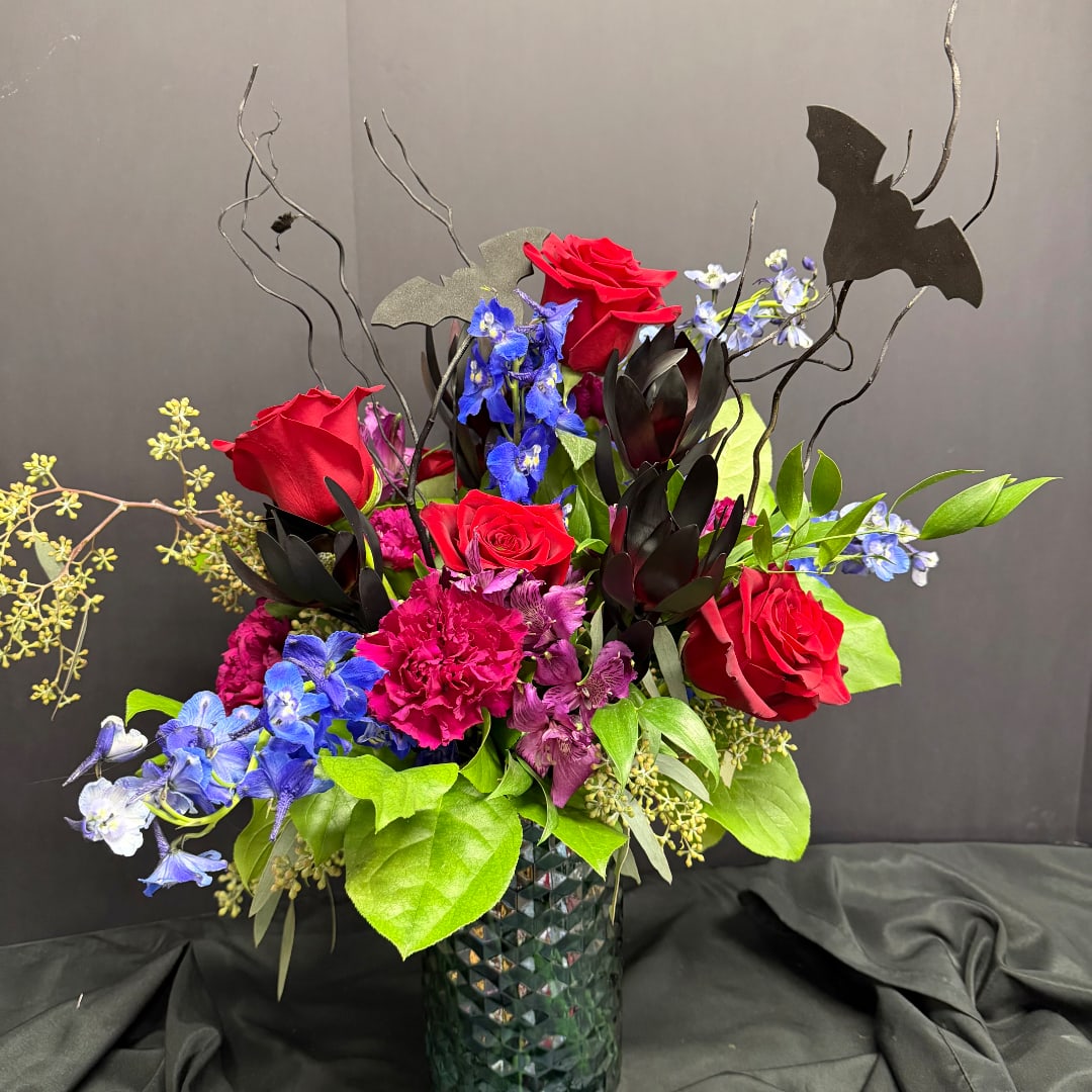 Red roses and blue flowers arranged in a dark vase with bat decorations.