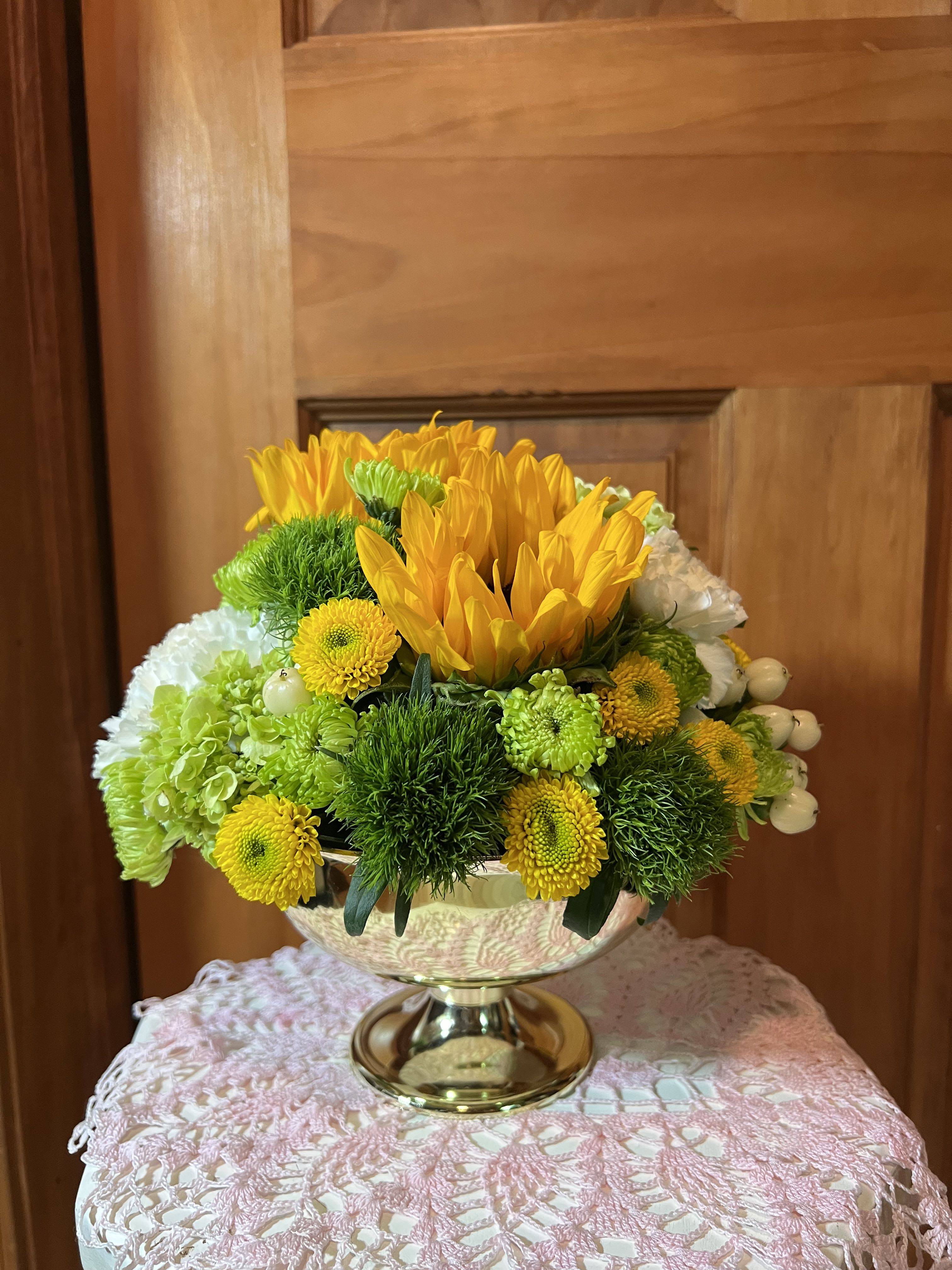 Sunny Days - Two tall sunflowers rise above a compact, rounded centerpiece-an upbeat flower arrangement perfect for a birthday, congratulations, or a sunny just-because gift. The mix of yellow chrysanthemum blooms with crisp mini green hydrangeas and lime-green button mums creates an immediate feeling of warmth and cheer that will make the person you're thinking of feel genuinely celebrated.  Arranged in a reflective gold pedestal bowl the design reads polished and slightly formal while staying fresh and spring-like. The composition is dense and low with lively textural contrasts-the plush hydrangea heads, bright daisy-like chrysanthemums, and tall sunflower faces-making it an ideal centerpiece for dining tables or reception counters.  Because of its compact, rounded shape this arrangement fits everyday displays and special gatherings alike; no elaborate care is needed beyond fresh water in the vessel. Choose this sunny arrangement when you want someone to feel noticed, uplifted, and truly seen.
