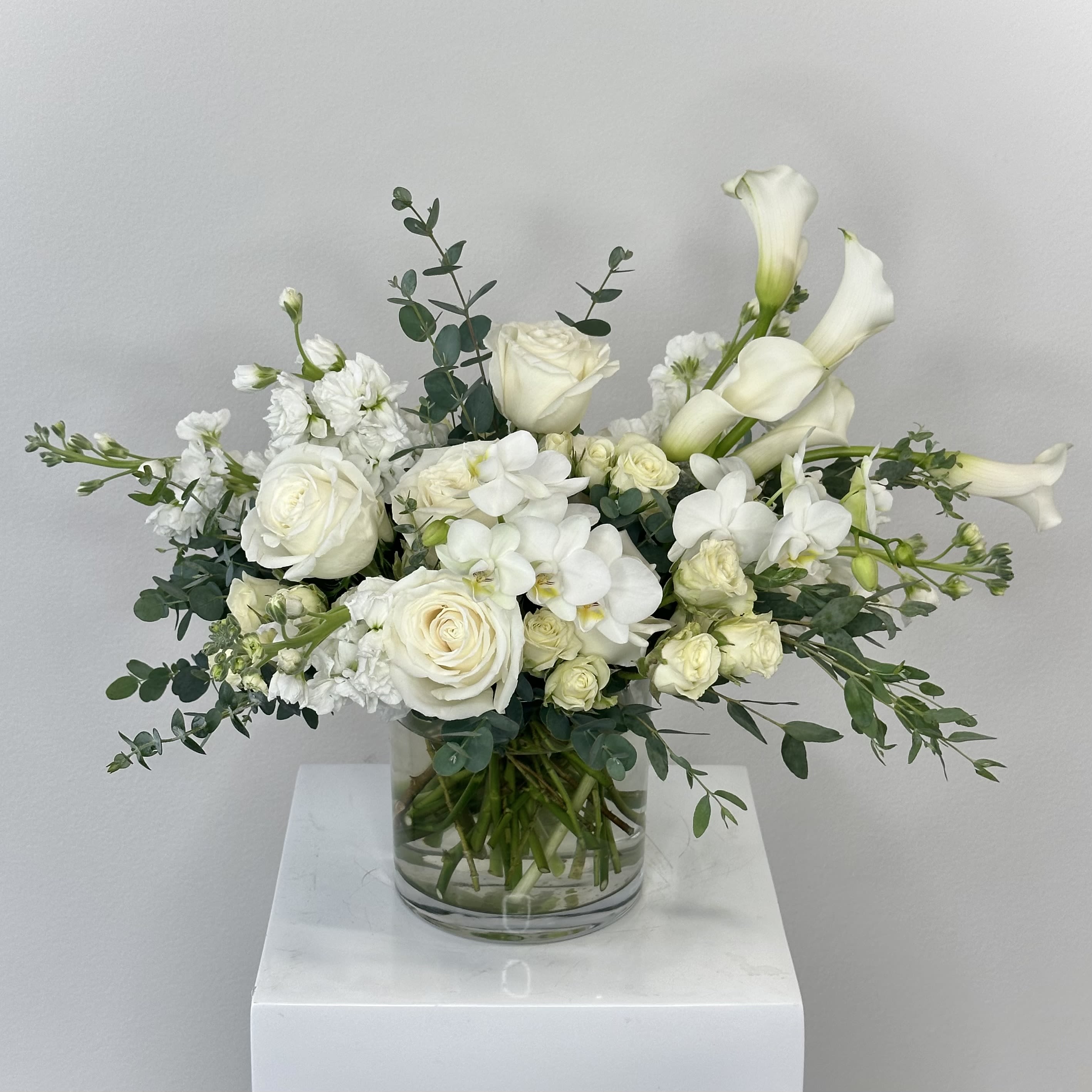 Purity - Discover the timeless elegance of Purity, a luxurious all-white flower arrangement from our parent company, Fleurs de Mad, that embodies innocence, serenity, and refined sophistication. This exquisite design showcases delicate mini white phalaenopsis orchids symbolizing purity and eternal love, paired with classic white roses and airy white spray roses for romance and grace, fragrant white stock adding height and sweet spice, sleek white calla lilies evoking beauty and rebirth, all accented by lush eucalyptus greenery for a fresh, natural touch and subtle herbal aroma. Handcrafted with premium fresh blooms in a monochromatic white palette, Purity creates a calming, ethereal aesthetic ideal for modern minimalist decor, elegant interiors, or serene spaces. The soft textures and varying forms—from the orchids' butterfly-like petals to the calla lilies' trumpet shapes—blend harmoniously with eucalyptus leaves for added depth and movement. Perfect as a breathtaking centerpiece for weddings, bridal showers, baptisms, sympathy tributes, or "thinking of you" gestures, this arrangement radiates peace and joy. Its symbolic white flowers make it a meaningful choice for celebrations of new beginnings, memorials, or simply elevating everyday luxury. Selected for exceptional freshness and longevity, Purity delivers enduring beauty and subtle fragrance. Bring tranquil sophistication to your home or gift this stunning all-white flower bouquet today!